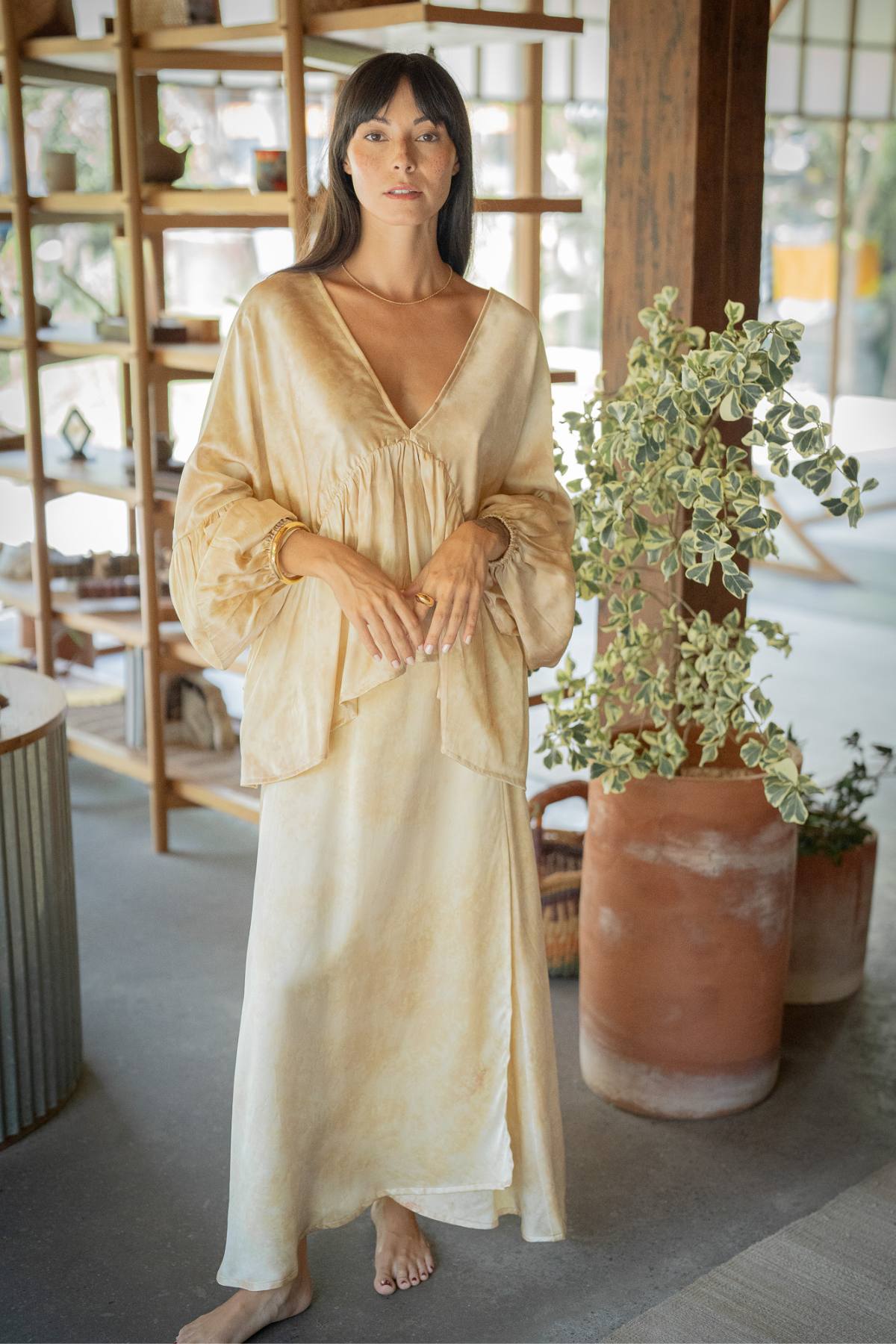 A woman with long dark hair stands barefoot, wearing the Eva Top & Shanti Skirt Set in a stylish light tone with billowy sleeves, crafted from 100% naturally dyed silk by Myrah Penaloza. She is in a room adorned with wooden shelves and various decorative items, including a leafy plant in a large pot. Natural light fills the space.