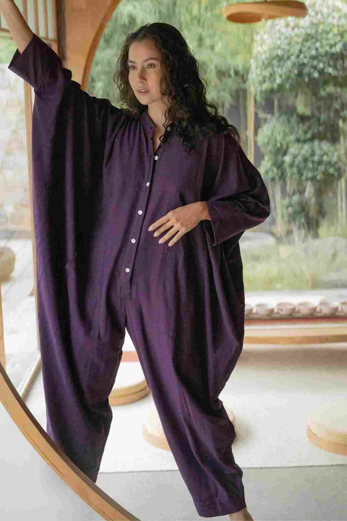 A person stands confidently inside a naturally lit room, wearing the flowing, deep purple Miracle Playsuit 2.0 from the Karama Collection by Myrah Penaloza. The playsuit features wide sleeves that enhance its elegant design. They have long, wavy hair and gracefully pose with one hand touching the curve of a wooden doorway while looking ahead. The serene green outdoor scene in the background complements their poised appearance.