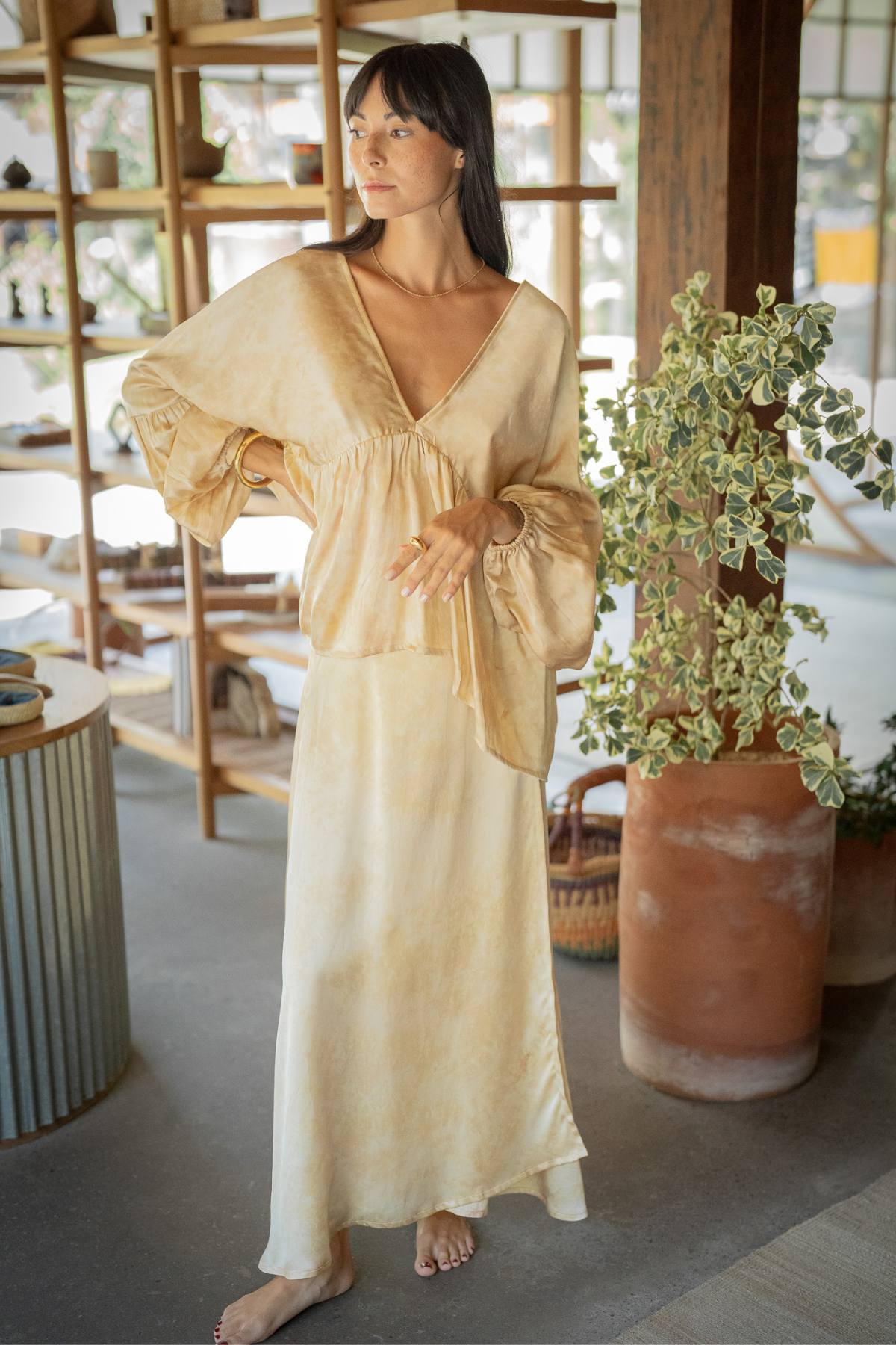 A person stands in a softly lit room wearing the "Eva Top & Shanti Skirt Set," crafted from 100% naturally dyed silk by Myrah Penaloza. They have long hair and are barefoot, with the background showcasing shelves with decor and a large potted plant.