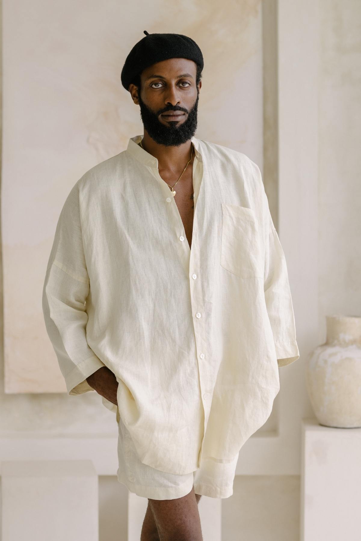 A man wearing the Camisa del Novio Mens Linen Top by Myrah Penaloza, paired with shorts and a black beret, stands in a neutral-toned room. He casually places his hands in his pockets as he gazes at the camera, framed by a large vase and a textured wall.