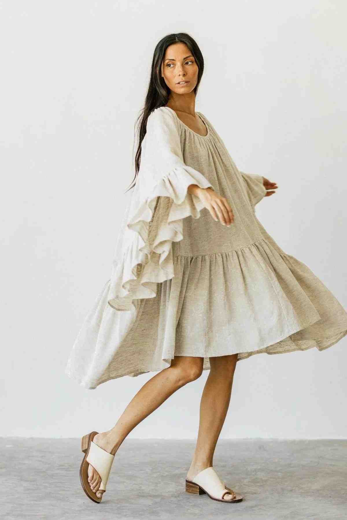 A person with long hair is wearing the Linen Brida Gown Short by Myrah Penaloza, a flowing and romantic dress perfect for spring and summer, featuring ruffled sleeves. They are twirling gracefully on a plain background, showcasing elegant white sandals.
