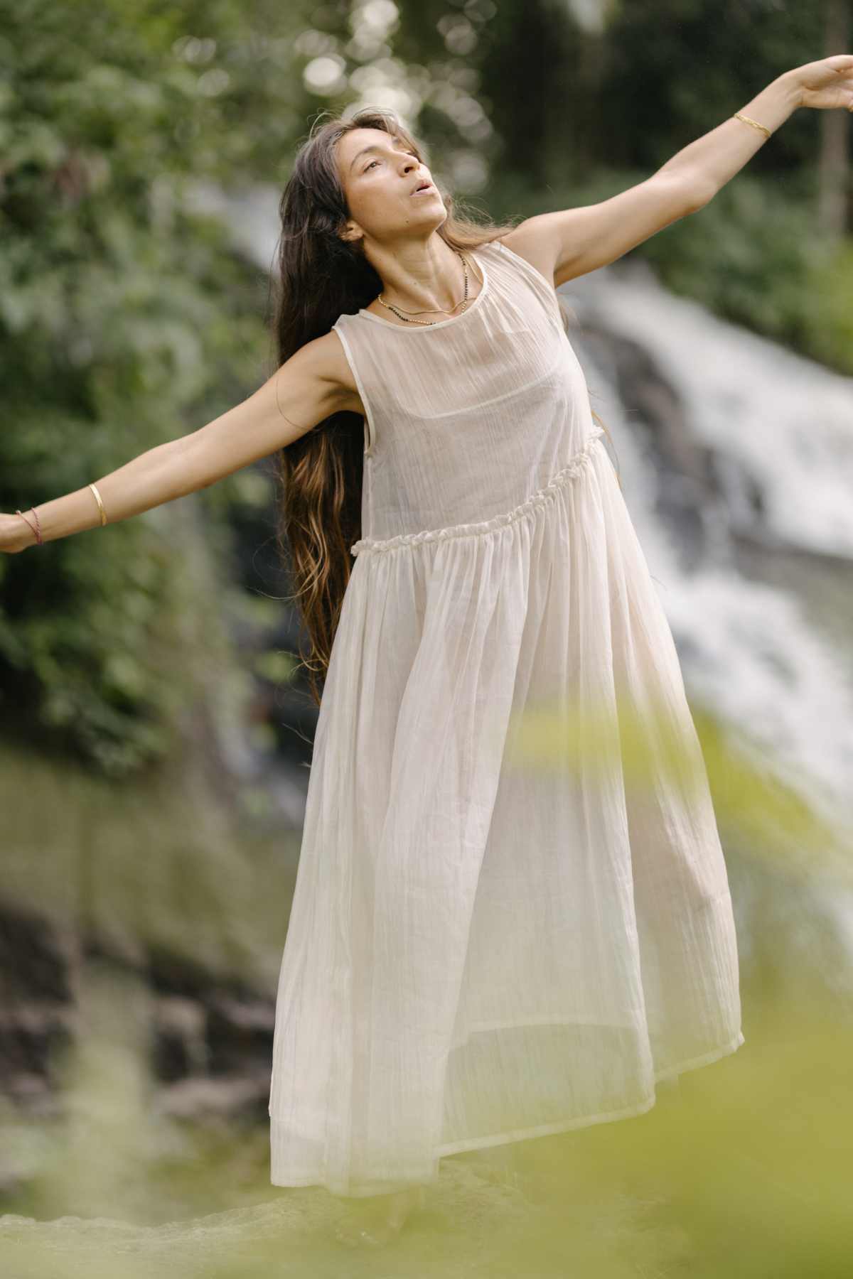 A woman, elegantly clad in a Dolly Night Gown (Sheer, 100% Chiffon Silk) from Myrah Penaloza and adorned with gold bangles, gracefully extends her arms while standing outdoors. She is enveloped by lush greenery with a waterfall cascading in the background, crafting a tranquil and natural ambiance.