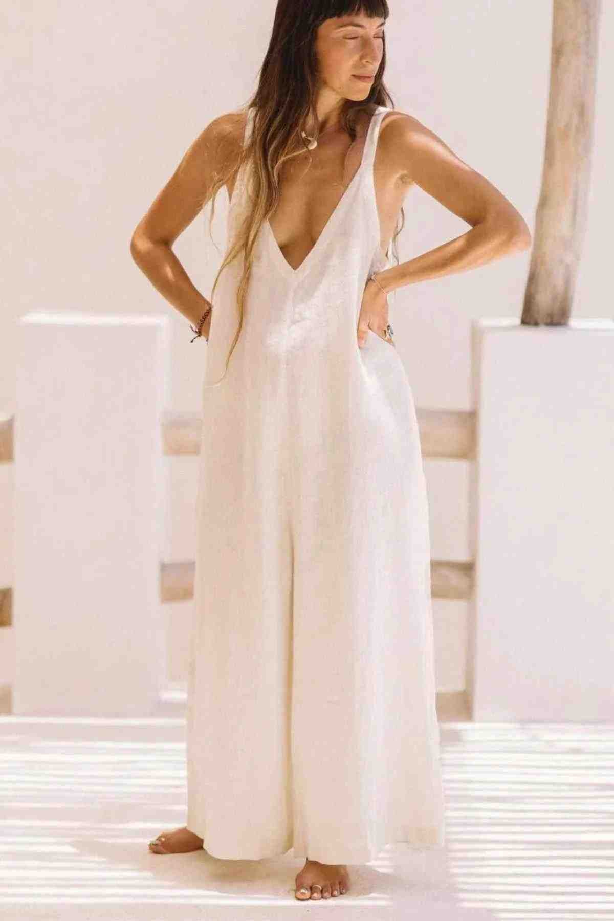 A woman stands with hands on hips, wearing the Kundalini Linen Playsuit by Myrah Penaloza—a flowing, sleeveless white jumpsuit featuring a deep V-neck and wide-leg bottoms. She has long, straight hair and is barefoot on a sunny day. The background is minimalistic with light-colored walls and a wooden post.