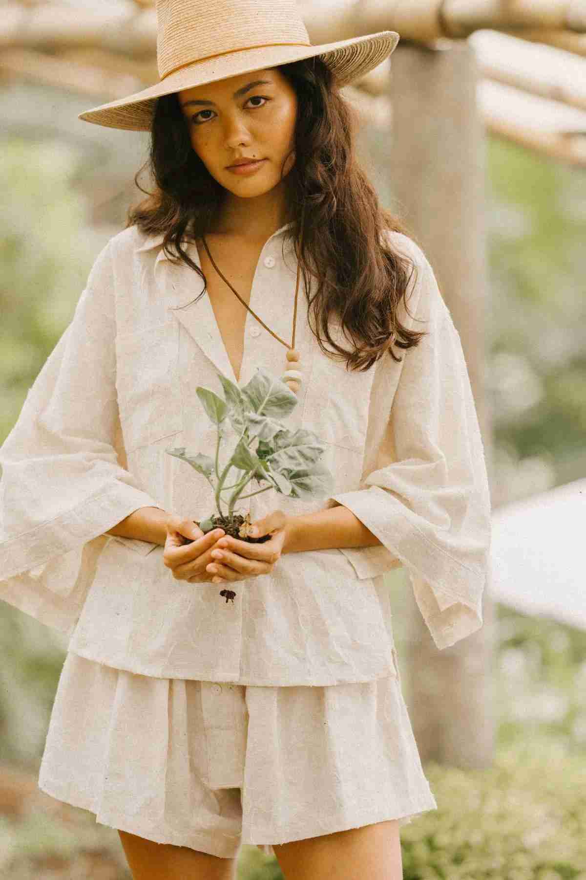 A person wearing the Linen Nidra Button Down Set from Myrah Penaloza and a straw hat stands outdoors, holding a small plant. The background of blurred greenery creates a serene and natural atmosphere.