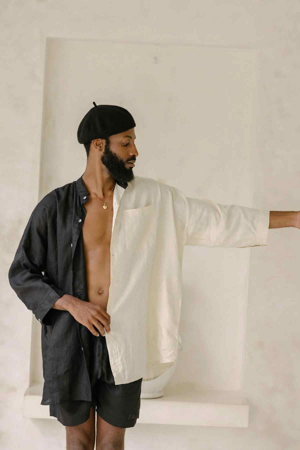 A person with a beard stands against a plain background, wearing the Unisex Yin Yang Linen Camisa Del Novio Set by Myrah Penaloza, which features a two-toned shirt with a black left side and white right side, reminiscent of the Yin Yang symbol. They are also sporting a black beret and black shorts, with their right arm extended to the side.
