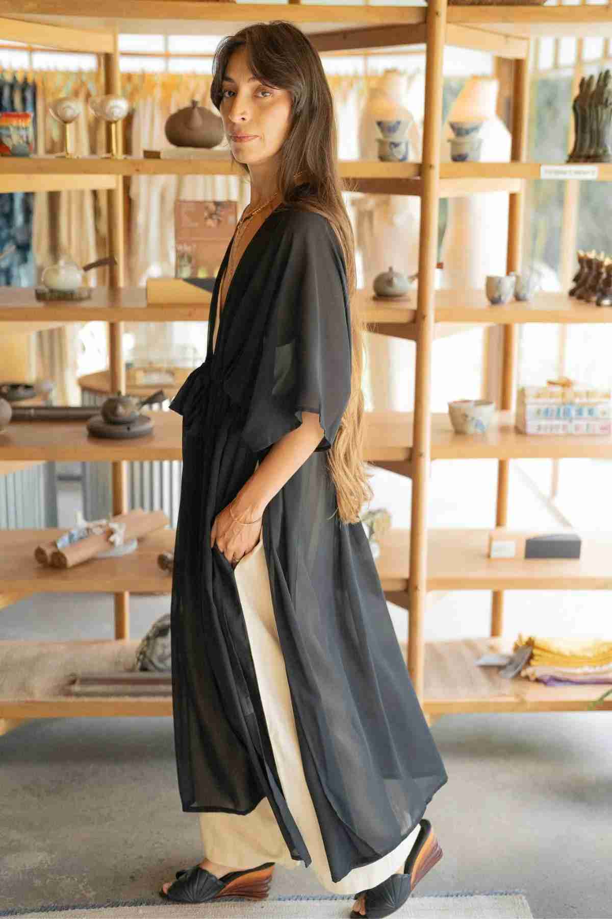 A woman with long hair is standing in front of a wooden shelf showcasing various items. She is adorned in a Madelin Sheer Chiffon Long Cover by Myrah Penaloza over light-colored pants and black sandals, radiating sheer fashion. The background features a brightly lit room with natural light illuminating the space.