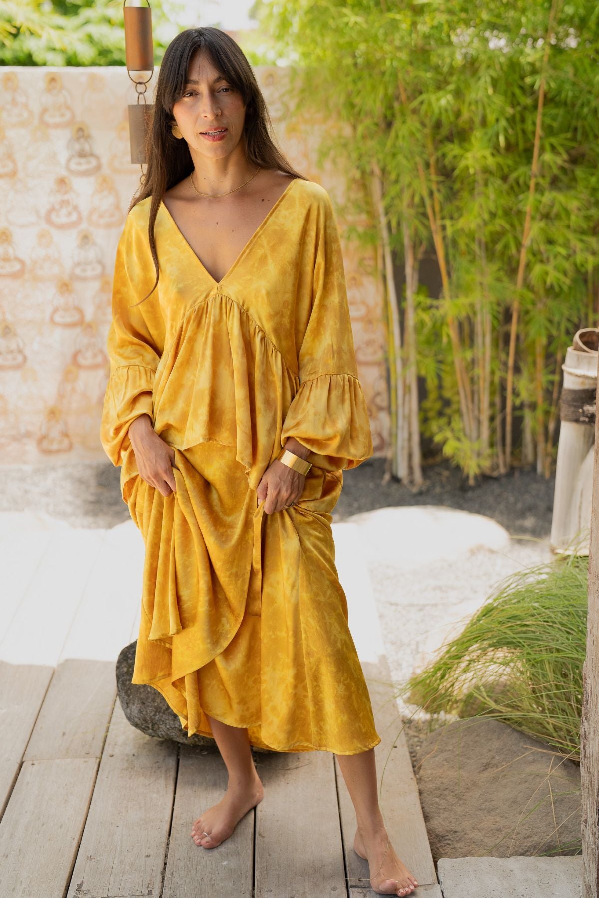 A person stands barefoot on a wooden deck, clad in the Myrah Penaloza Eva Top & Shanti Skirt set, crafted from 100% naturally dyed silk. They are outdoors, surrounded by bamboo plants and soft lighting, conveying a peaceful, bohemian atmosphere.