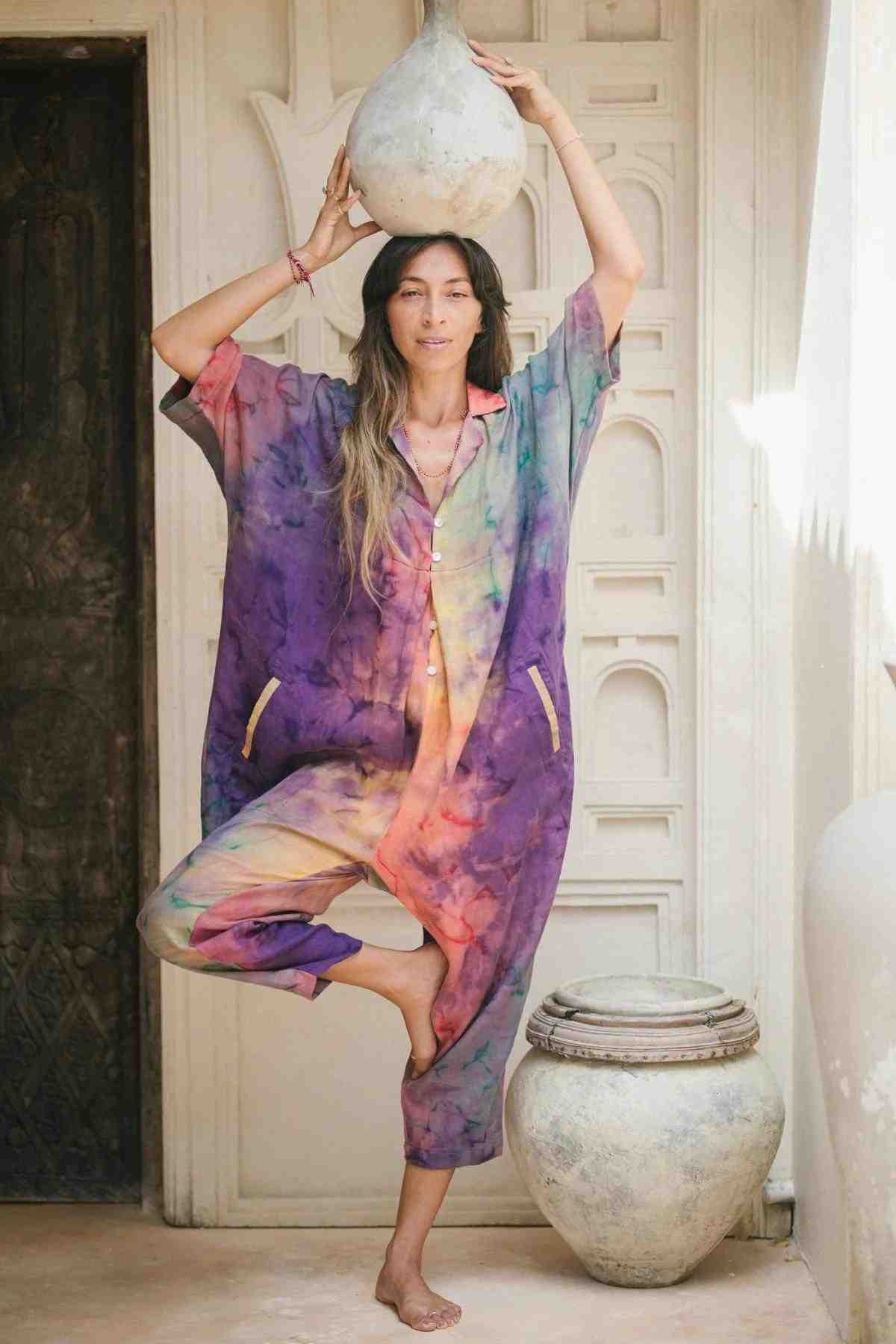 A woman stands in a yoga pose on one foot, balancing a large ceramic pot on her head. She is dressed in the colorful, tie-dye Sat Torri Rainbeau Playsuit by Myrah Penaloza and has long hair. She poses against a textured wall with another ceramic pot placed near her feet. Sunlight filters into the scene, evoking hints of retro 1980s design.