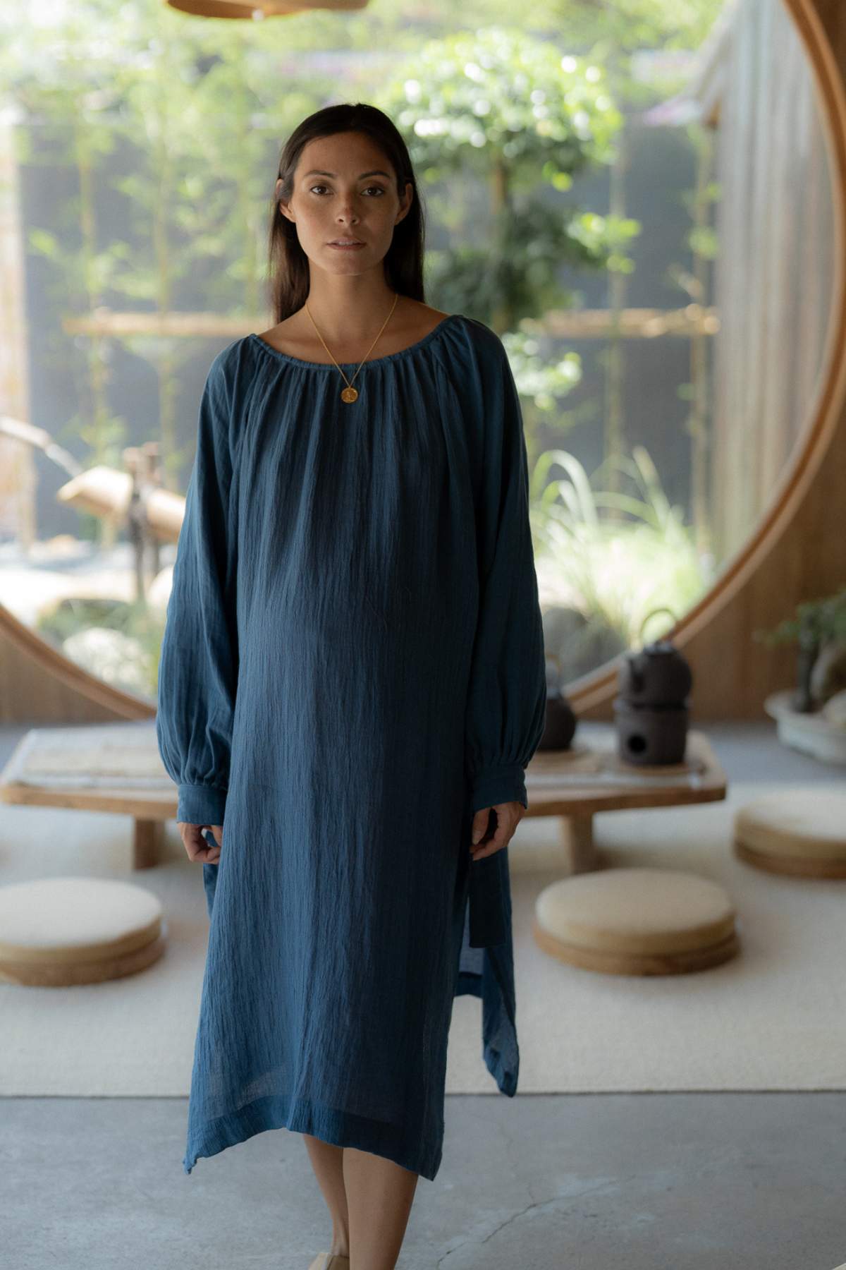 A woman wearing the reversible Longing Gown by Myrah Penaloza stands in a serene, minimalist room bathed in natural light. The space, with its circular windows, low seating, and green plants, exudes a tranquil atmosphere.