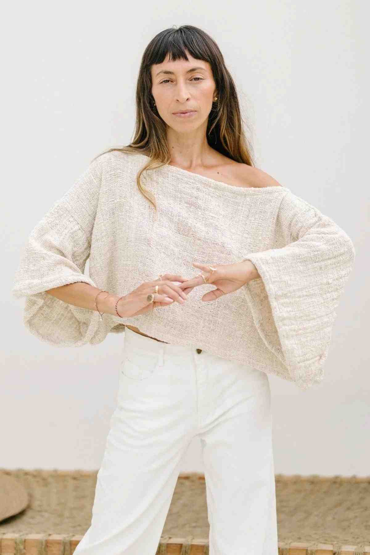 A woman with long, dark hair and bangs is standing with her eyes closed. She is wearing an oversized, light, loose-fitting, off-the-shoulder Tumanggal Cotton Spirit Top by Myrah Penaloza with flared sleeves and white pants. She has several minimalistic rings and bangles on her hands. The background is plain and neutral.