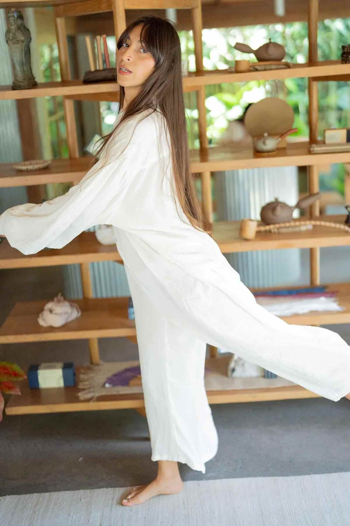 A person with long hair wearing a flowing white Kundalini Gown Playsuit Edition (Pre-Order) from Myrah Penaloza is standing on one leg in front of a wooden shelf filled with various items. The background shows greenery outside through a large window. The person appears to be balancing or performing a light dance move.