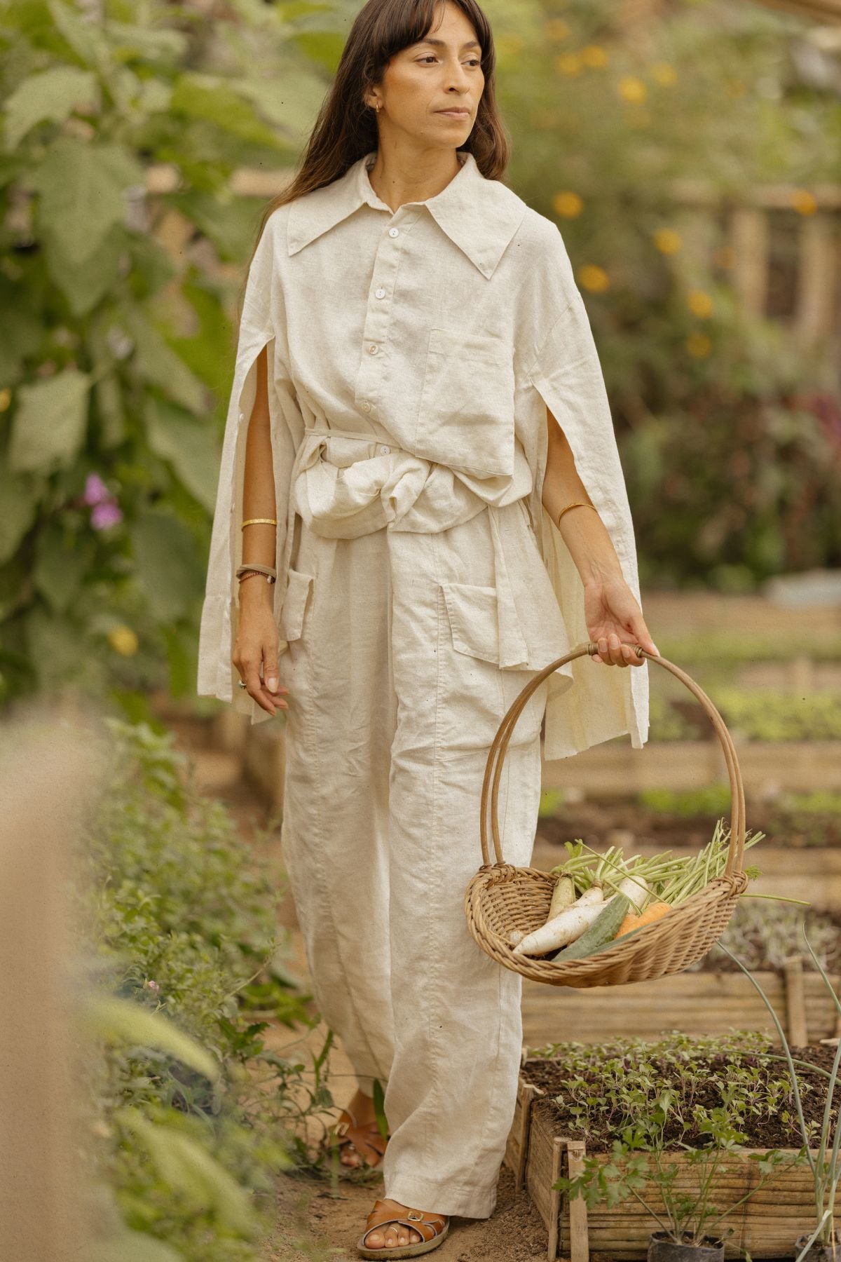 A woman wearing the light beige and flowing Sutra Linen Set from Myrah Penaloza stands in a garden, holding a wicker basket brimming with freshly picked vegetables like carrots and beans. The background showcases lush green plants and raised garden beds. She gazes thoughtfully into the distance, epitomizing versatile elegance.