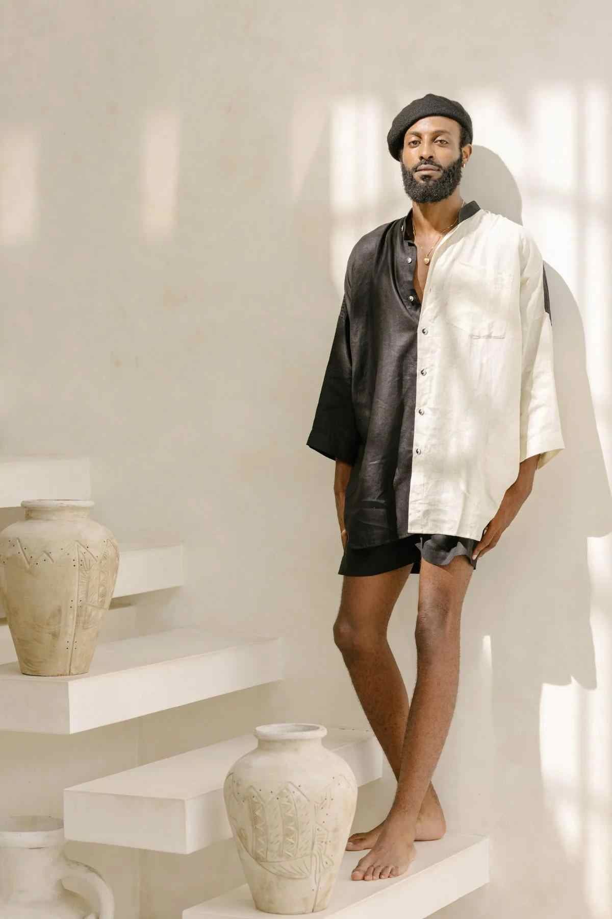 A tall individual with a beard stands on a white staircase next to large ceramic pots. They are dressed in the Myrah Penaloza Unisex Yin Yang Linen Camisa Del Novio Set, which includes a loose-fitting, two-tone shirt (black on one side and white on the other) adorned with a Yin Yang linen patch, black shorts, and a black beret. The background is minimalist and bathed in natural light.