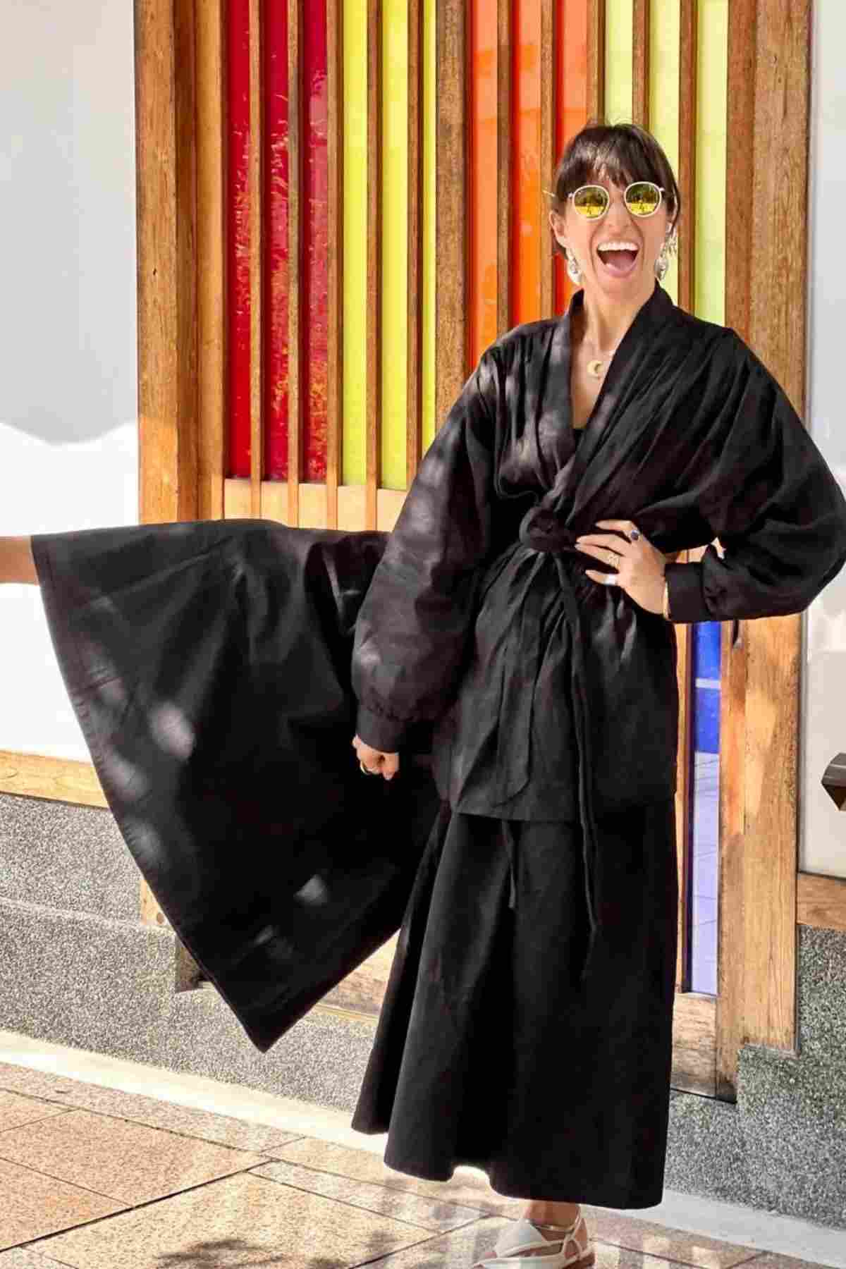 Dressed in a flowing black outfit and round sunglasses, a person joyfully poses with one hand on their hip, showcasing the Pantalon Linen Pants (Wide Legged) from Myrah Penaloza as they stand before a vibrant, striped glass window.