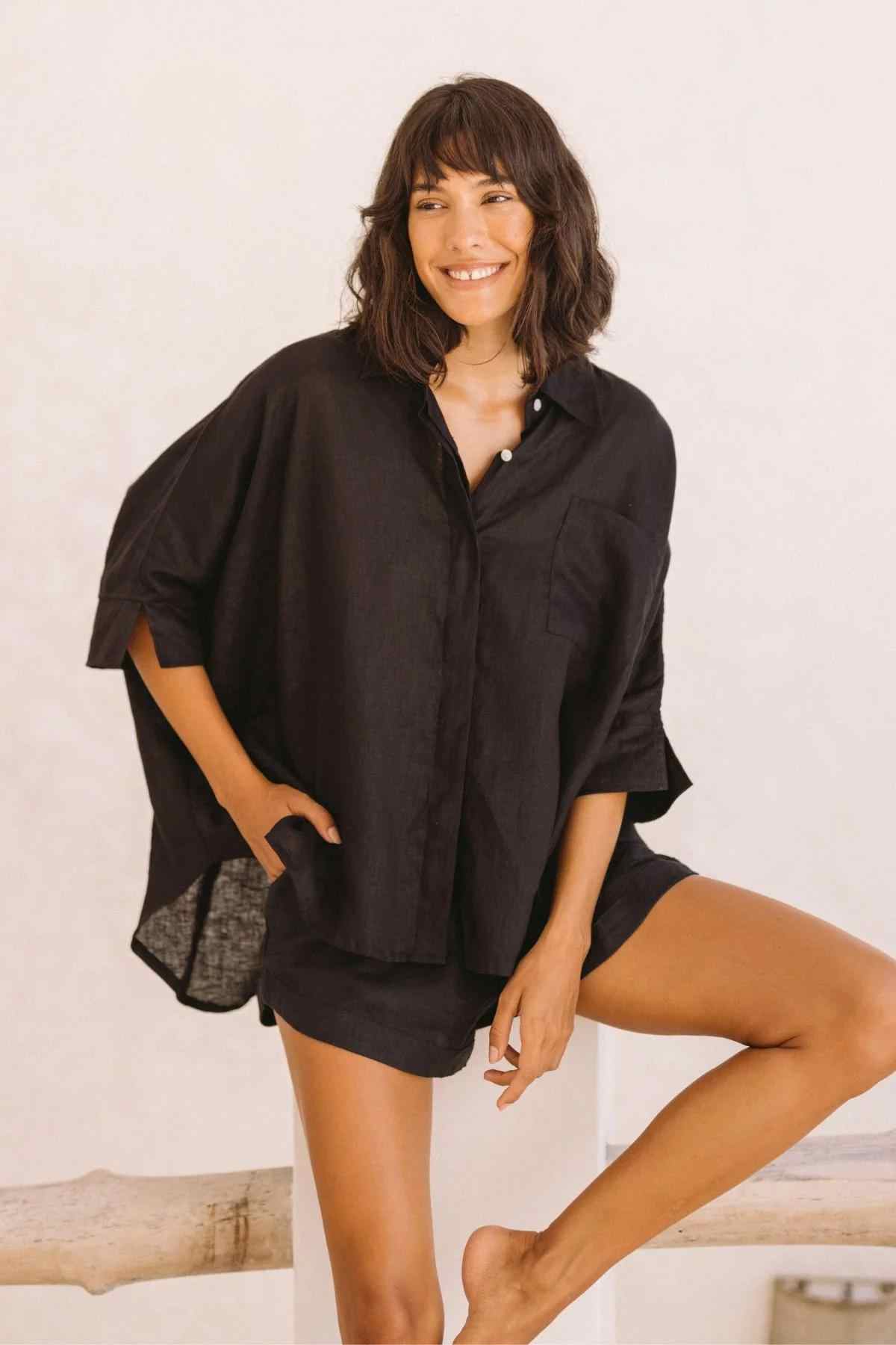 A woman with shoulder-length dark hair is smiling while leaning against a white surface. She is rocking the Myrah Penaloza Linen Suka Button Down Set, a loose-fitting black shirt and matching shorts made from sustainable linen. One hand rests on her hip, and the other is on her thigh. The background is plain and light-colored.