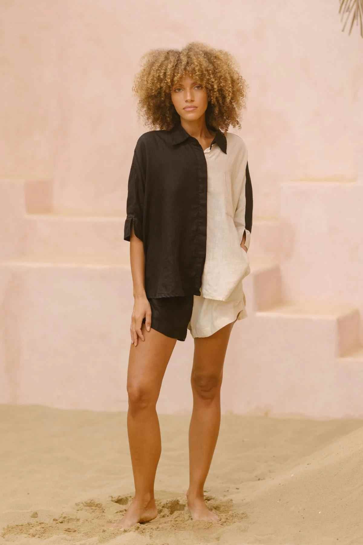 A woman with curly hair stands barefoot on sand, adorned in the Yin Yang Suka Set from Myrah Penaloza. The soft pink steps and walls behind her create a warm, minimalistic setting. With a relaxed poise, she gazes forward, embodying the essence of ethical fashion.