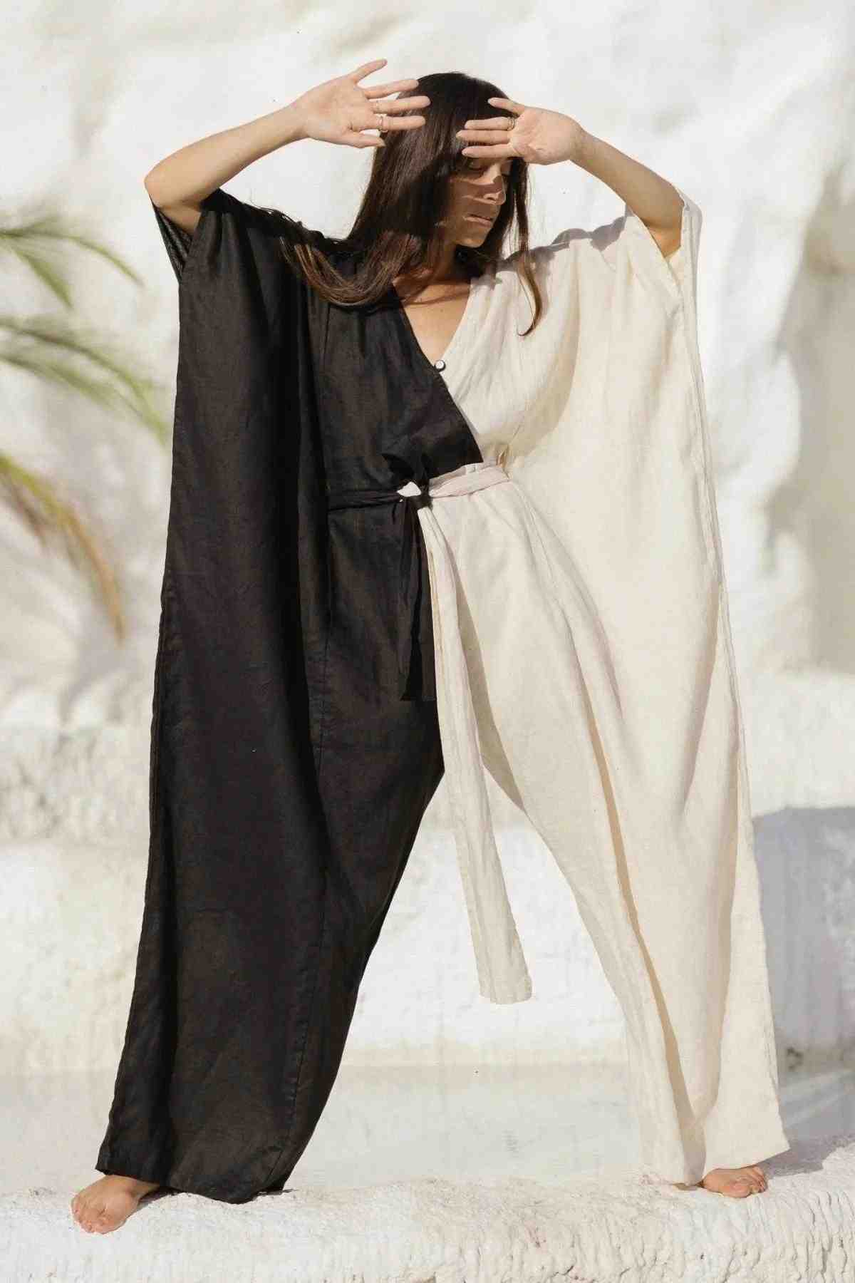 A person standing outdoors, elegantly dressed in the French Linen Yin Yang River Playsuit by Myrah Penaloza, featuring a split-colored flowing design with one side black and the other side white. The individual, with long hair cascading down, strikes a pose with arms raised and wrists crossed near their forehead against a textured light-colored background.