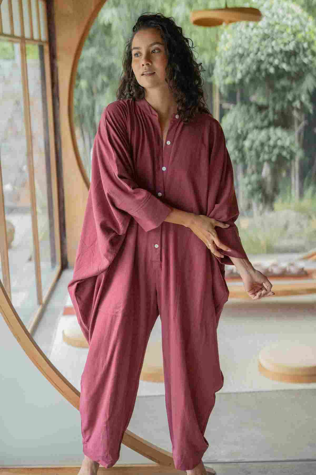 A person with curly hair stands in front of a large, circular window, wearing a loose-fitting, maroon Miracle Playsuit 2.0 from Myrah Penaloza's Karama Collection. They have a relaxed, contemplative expression, with one hand resting on their opposite arm. Natural scenery is visible outside.