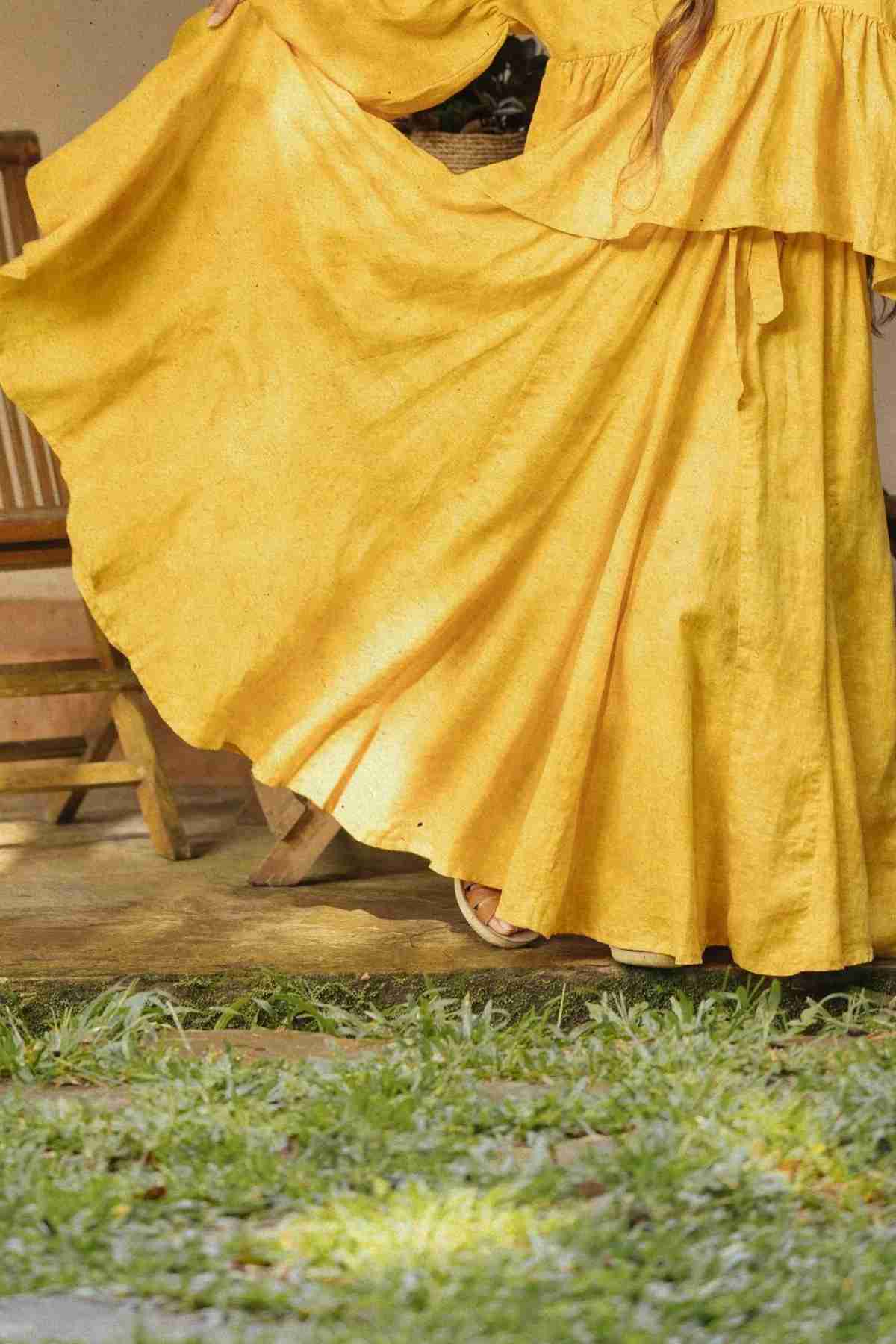 A person wearing a flowing, bright yellow Orisa Dance Skirt (100% linen) by Myrah Penaloza stands with their arms extended, causing the skirt to fan out gracefully. The background features an old wooden chair and part of a wooden structure, with green grass in the foreground.