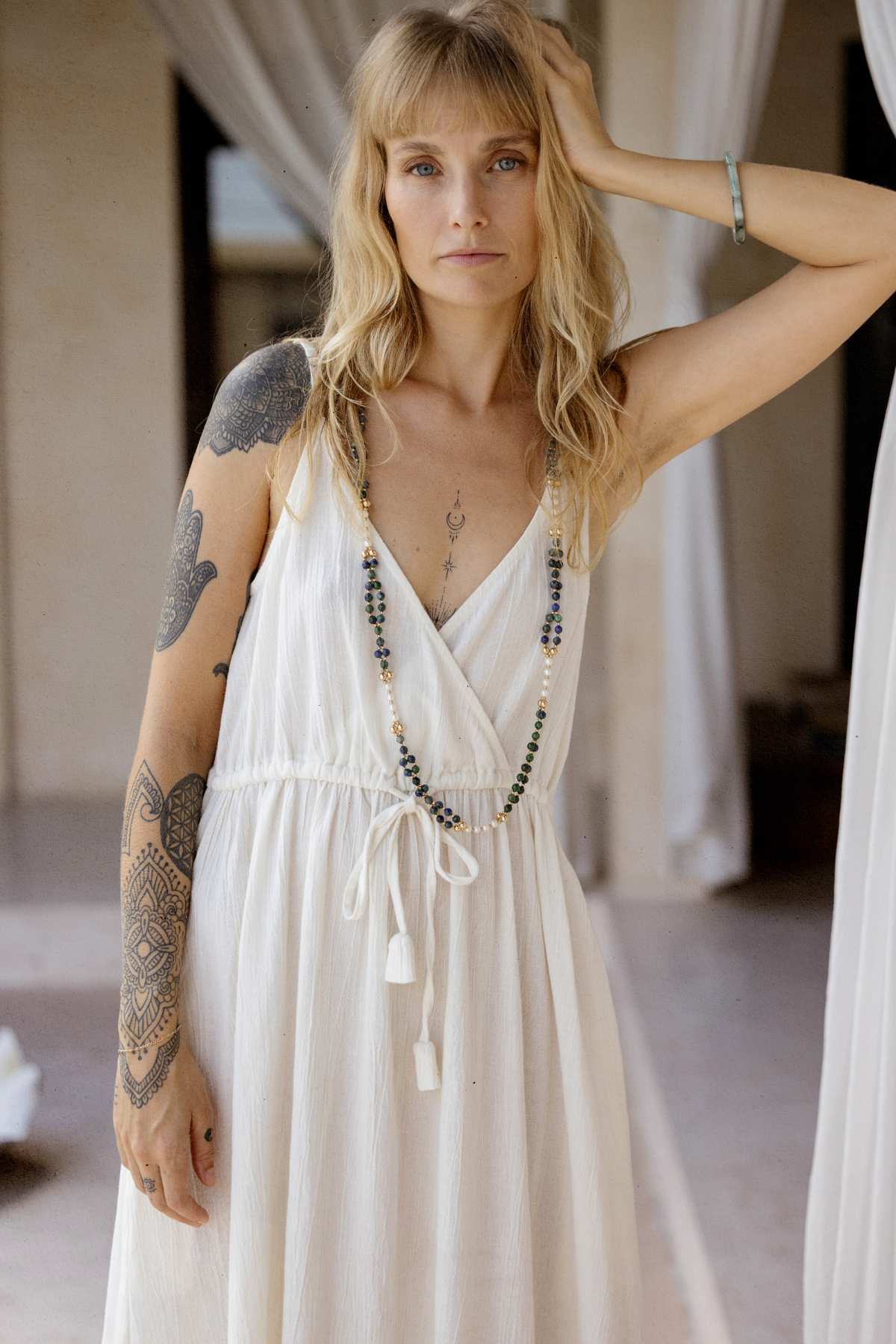A woman with long blonde hair and tattoos exudes effortless elegance in a sleeveless white Dora Slip Dress by Myrah Penaloza, complemented by a long bead necklace. She stands in a relaxed pose indoors, bathed in soft natural light with curtain drapes framing this timeless ensemble.