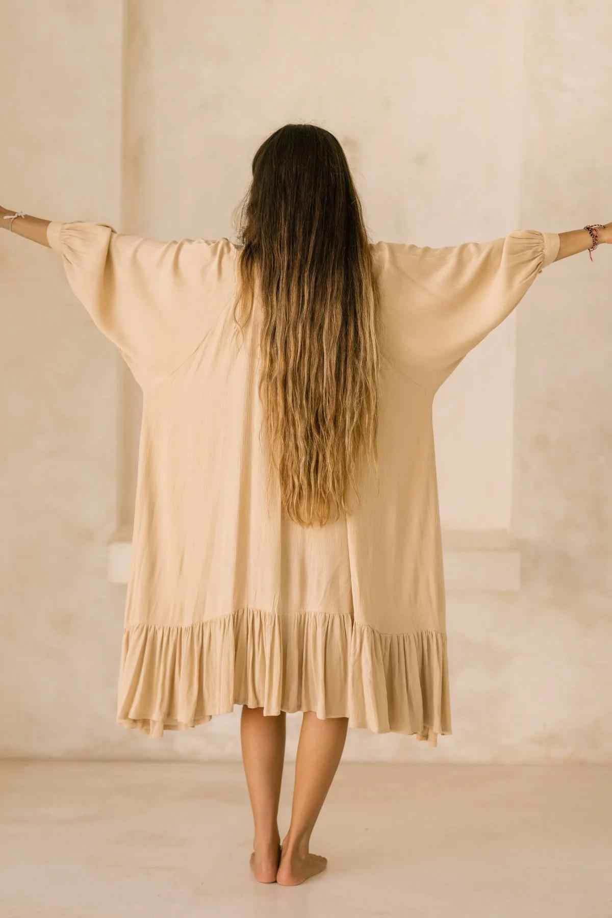A woman with long wavy hair stands barefoot in a flowing bamboo rayon gown from Myrah Penaloza, similar to the FRIDA Gown Short. Her arms are outstretched as she faces a light-colored wall. The beige dress is highlighted by its loose sleeves and ruffled hem.