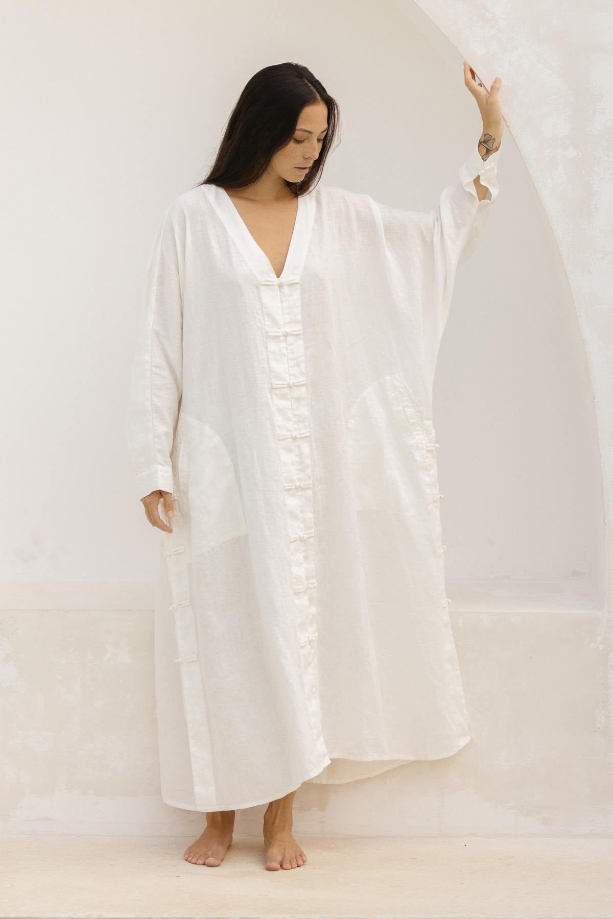 A barefoot woman with long dark hair stands against a pale wall, adorned in Myrah Penaloza's Ancient Chajin Jacket, a loose and flowing white dress crafted from 100% linen and made to order. Featuring a deep V-neck and large sewn-on pockets, the dress showcases delicate Japanese-inspired buttons that add an elegant touch to the ensemble. One arm is raised, lightly touching an arch as she gazes downward.