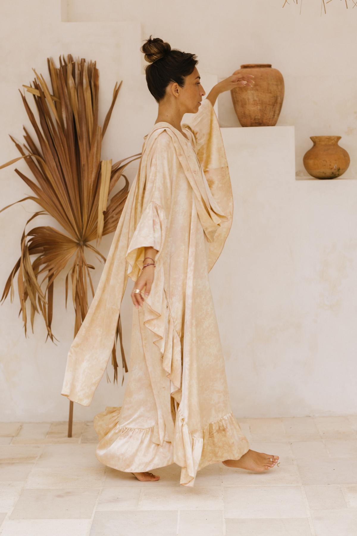 A woman with her hair in a bun stands in profile, wearing the Perla Silk Gown Made To Order by Myrah Penaloza. She is indoors, near a white staircase adorned with three terracotta pots on different steps. A dried palm leaf decorates the wall in the background—creating a scene reminiscent of a Florence and The Machine album cover.