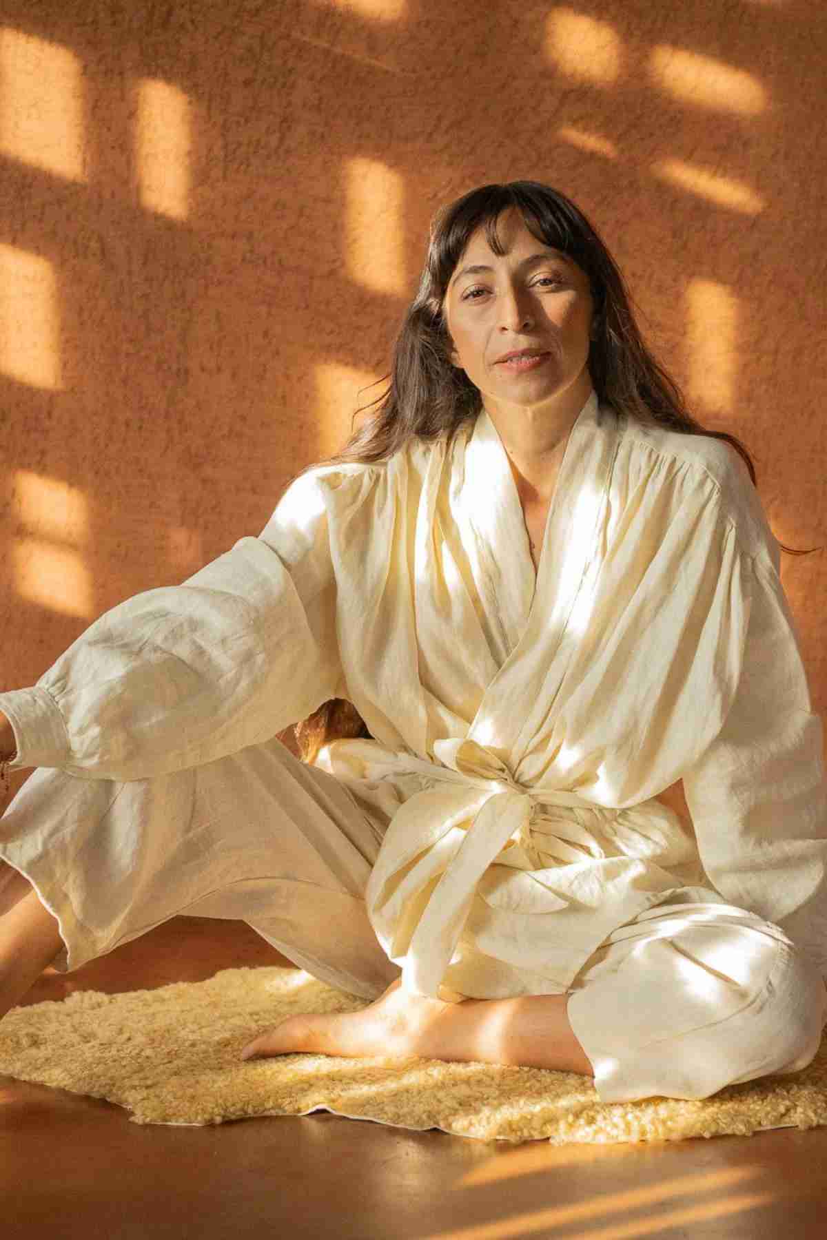A person with long, dark hair sits cross-legged on a soft surface, wearing the Linen Jasmine Set by Myrah Penaloza, a light and kimono-inspired outfit. The background features a warm, sunlit pattern on the wall. The person has a serene expression and is bathed in natural light.