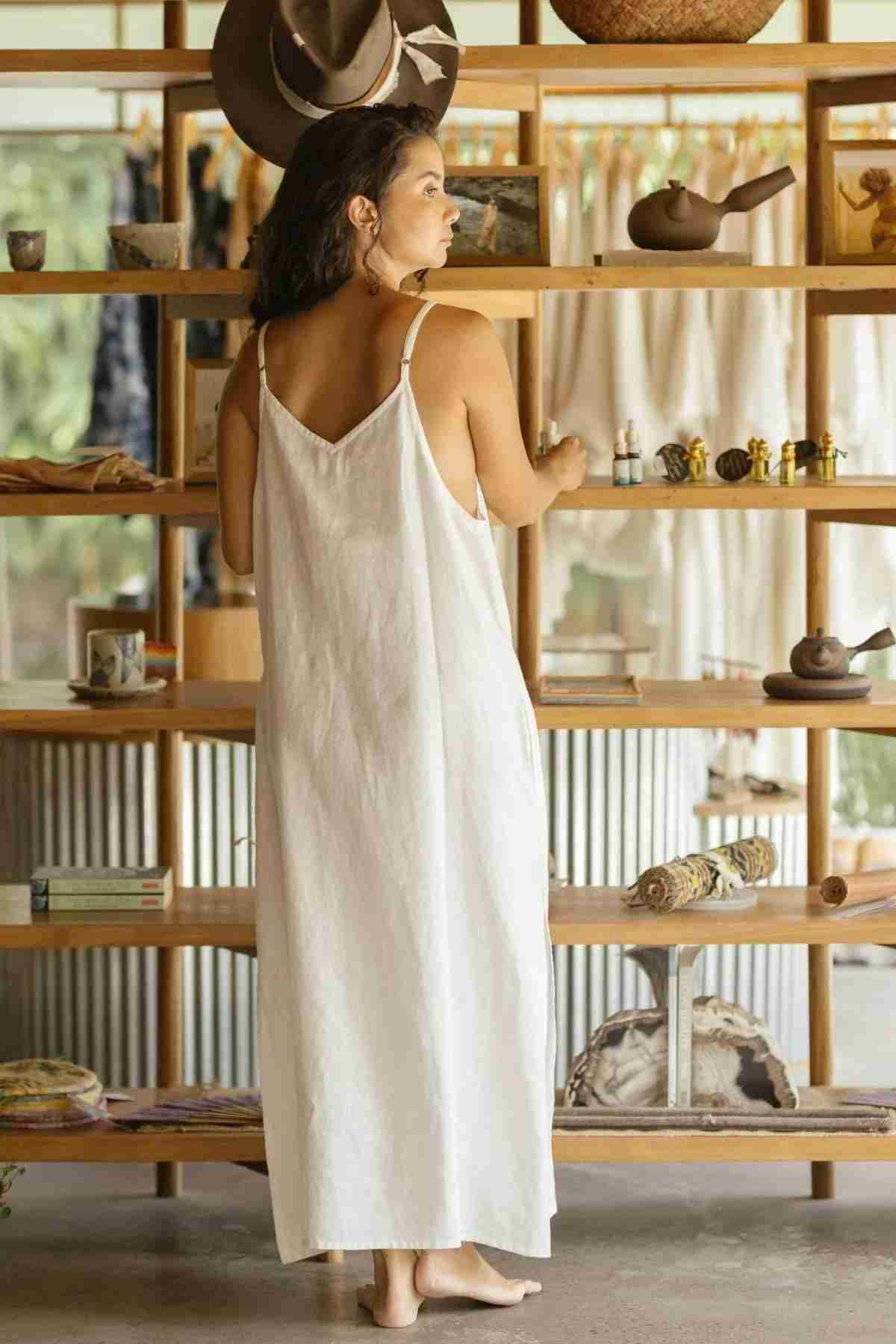 A woman in the Everyday Linen Slip by Myrah Penaloza stands barefoot in front of a wooden shelf display, admiring various decorative items, ceramics, and textiles. The background features a cozy interior with natural light, wooden floors, and a rustic ambiance that exudes luxurious comfort.