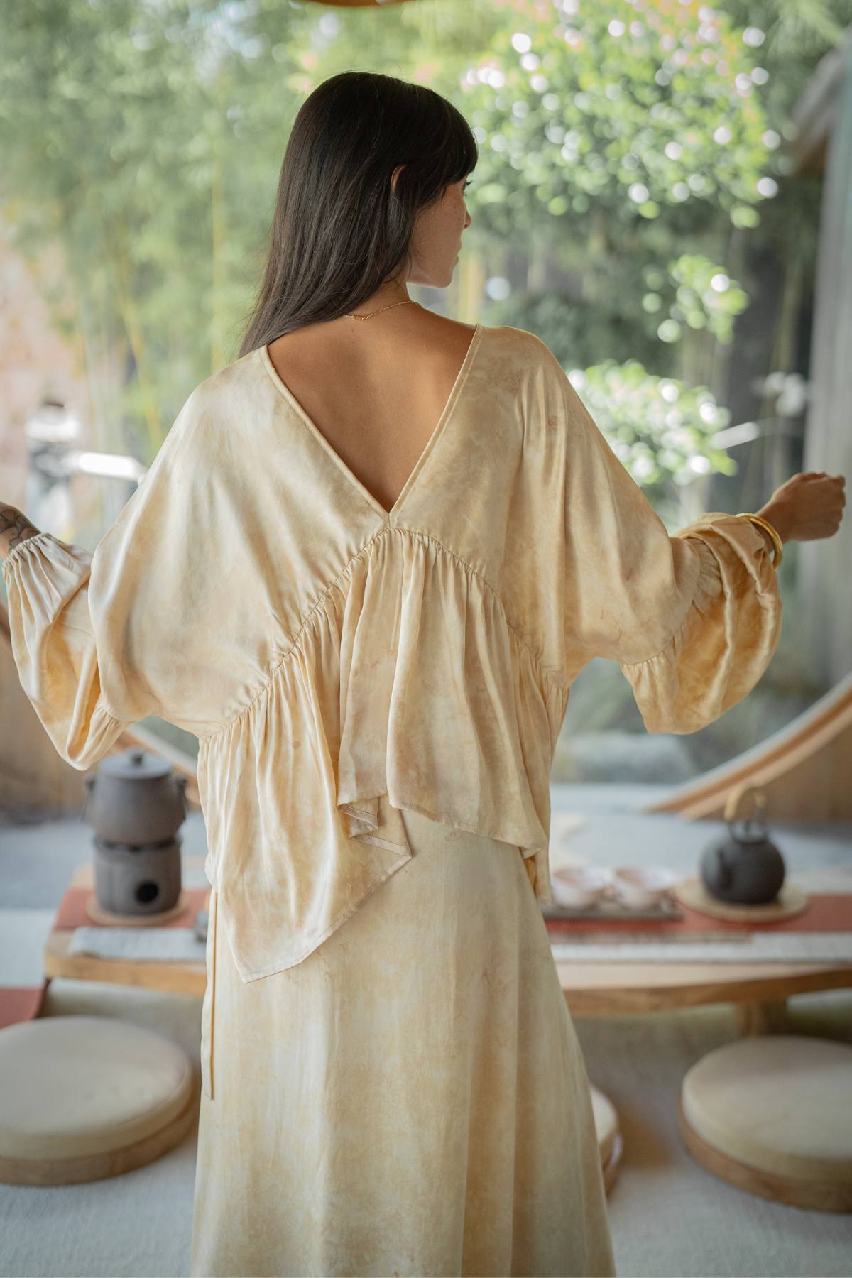 A person with long dark hair is wearing the flowy Myrah Penaloza Eva Top & Shanti Skirt Set, crafted from 100% naturally dyed silk. They stand in a softly lit room, surrounded by round wooden cushions and pottery, gazing out towards the garden.