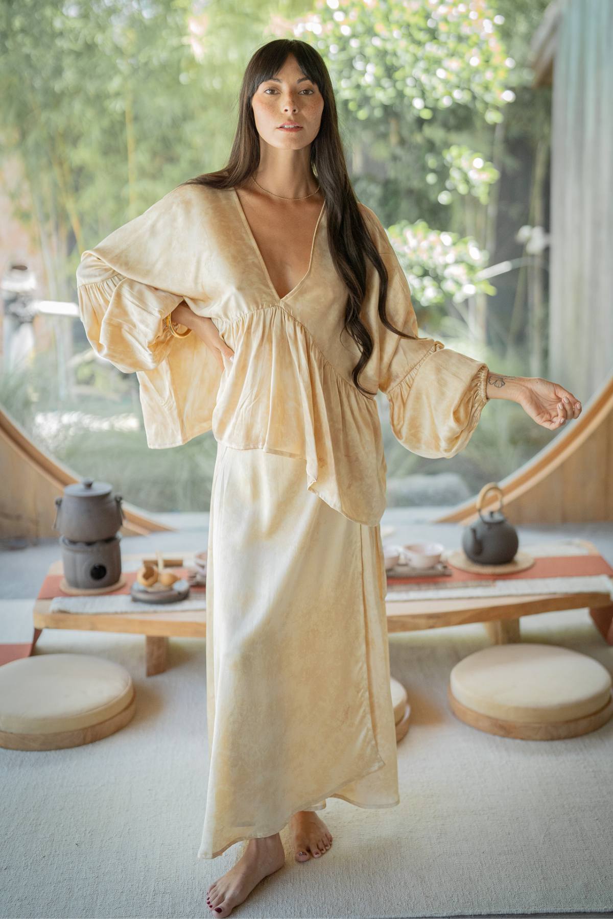 A woman with long dark hair is barefoot in a peaceful indoor setting, wearing the Eva Top & Shanti Skirt Set by Myrah Penaloza. This attire, crafted from 100% naturally dyed silk, flows gracefully as she stands near a wooden table adorned with a tea set. Soft natural light fills the room, enhancing the tranquil atmosphere with her sustainable fashion choice.