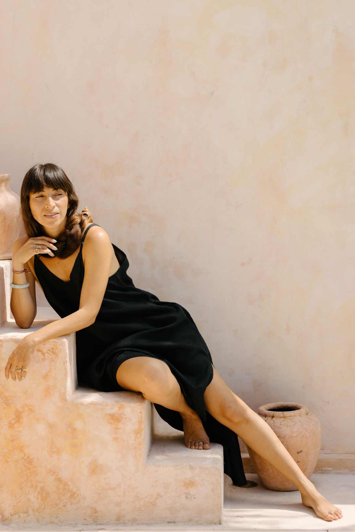 A woman with shoulder-length dark hair is seated on an outdoor staircase, elegantly adorned in the Everyday Linen Slip by Myrah Penaloza. Her sleeveless black dress exudes luxurious comfort as she rests one hand on the step and the other on her chin. The backdrop of a beige plastered wall and a clay pot adds to the scene's elegant yet relaxed ambiance.