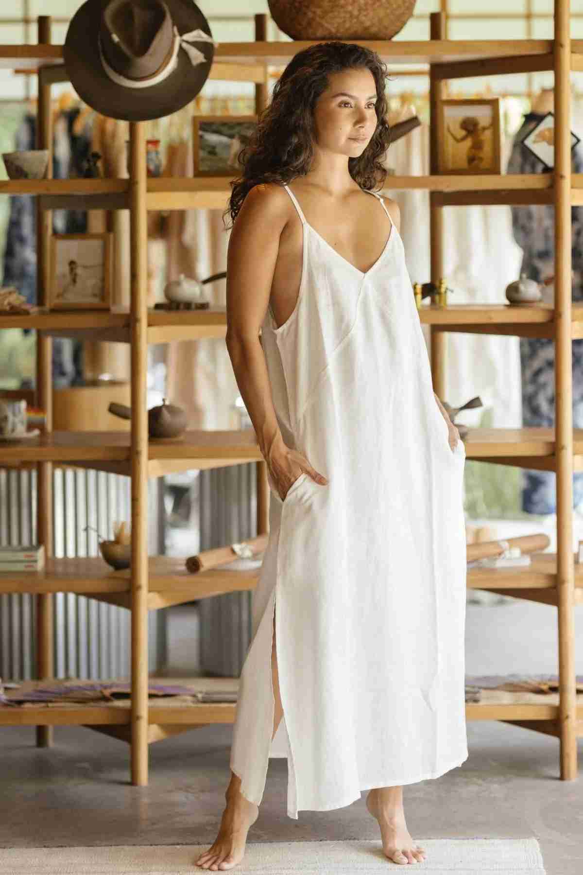 A woman with long curly hair stands barefoot on a light-colored rug in a room adorned with wooden shelves and various items. She is wearing the Everyday Linen Slip by Myrah Penaloza, a loose, sleeveless white dress made of stone washed linen, featuring side splits and luxurious comfort. A brown hat hangs on a shelf behind her.