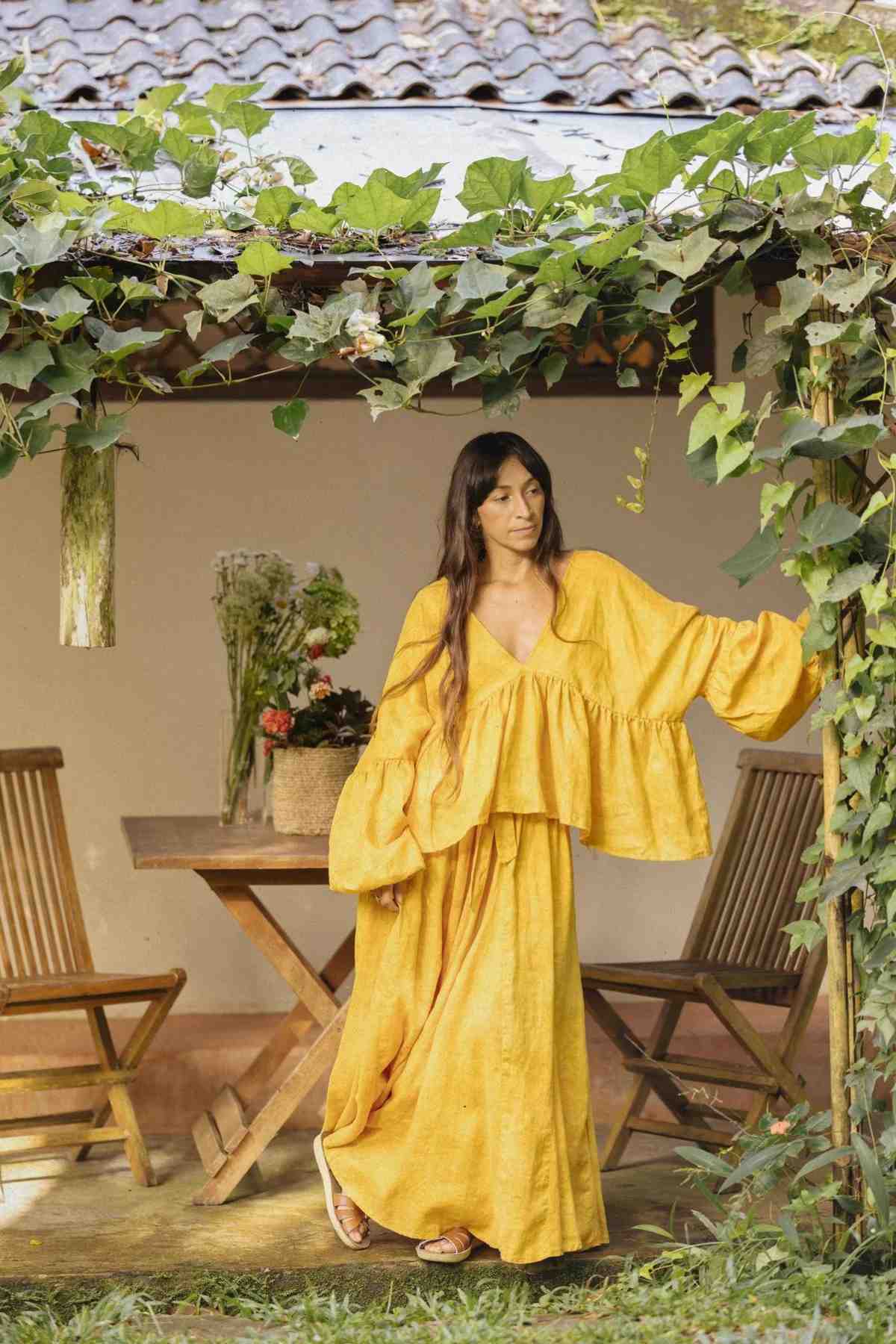 A woman with long dark hair stands under a vine-covered roof, wearing the flowing Linen Eva Top & Orisa Dance Skirt Set by Myrah Penaloza, its billowy sleeves catching the light. She leans against a wooden post while looking to the side. The serene scene includes a table adorned with a basket of flowers and two wooden chairs, hinting at sustainable linen decor.