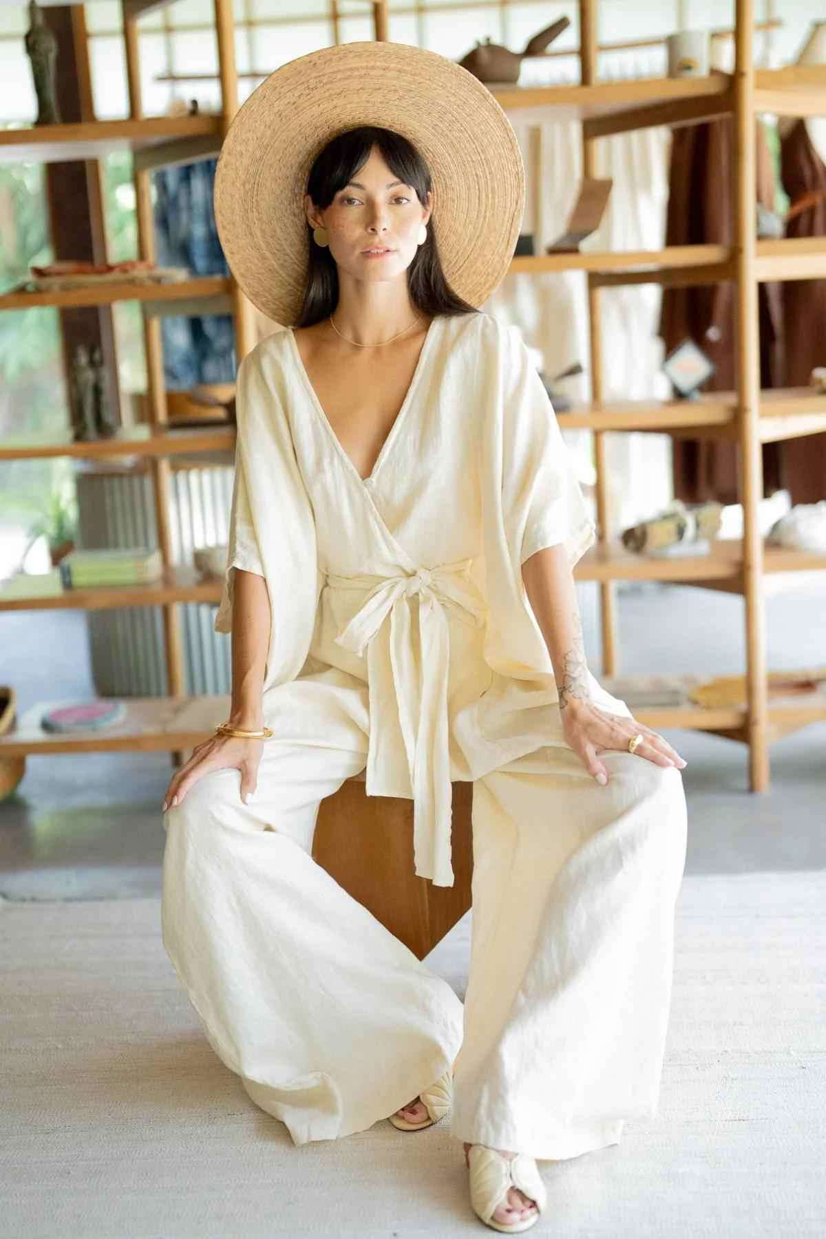 A woman sits indoors, adorned in a wide-brimmed hat and a Loose, light-colored Linen River Playsuit from Myrah Penaloza. Surrounded by wooden shelves with various items, she seems to be at a boutique showcasing collections including the River Kaftan along with stone-washed linen wear perfect for expectant mothers.