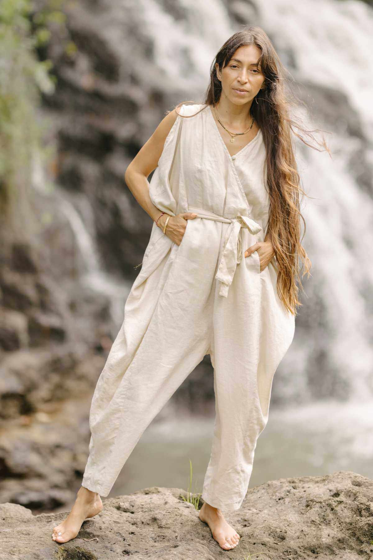 A woman stands barefoot on a rock with her hands in her pockets, wearing the loose, beige French Linen Kuan Yin Playsuit by Myrah Penaloza, which features an adjustable waist tie. She has long, wavy hair and is posing in front of a cascading waterfall. The natural background includes rocks and greenery.