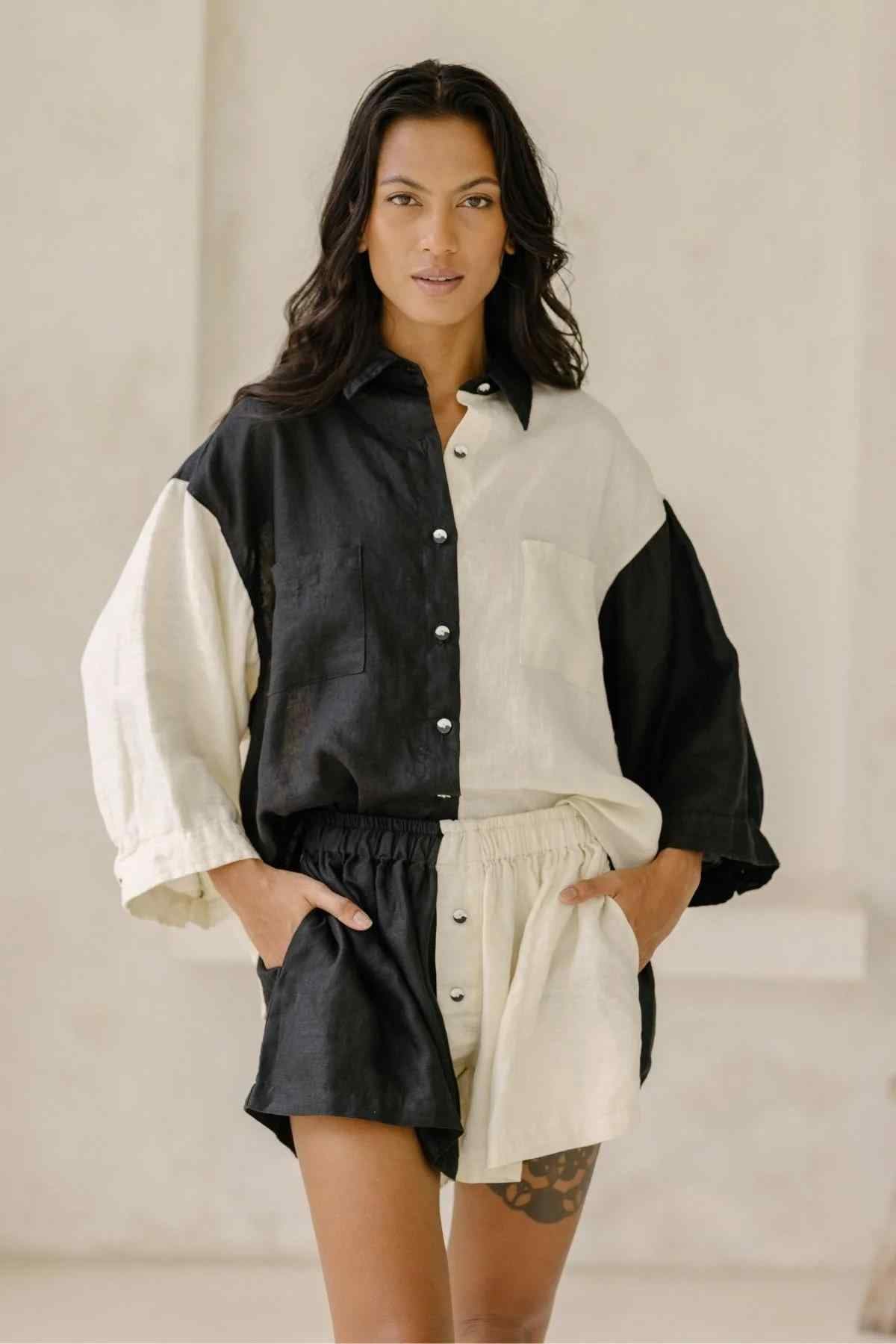 A person stands confidently with hands in pockets, modeling the stylish Yin Yang Nidra Button Down Set from Myrah Penaloza. The black and white color-blocked linen button-down shirt and matching shorts feature a relaxed silhouette. The individual’s long dark hair and neutral expression complete the look against a plain, light-colored background.