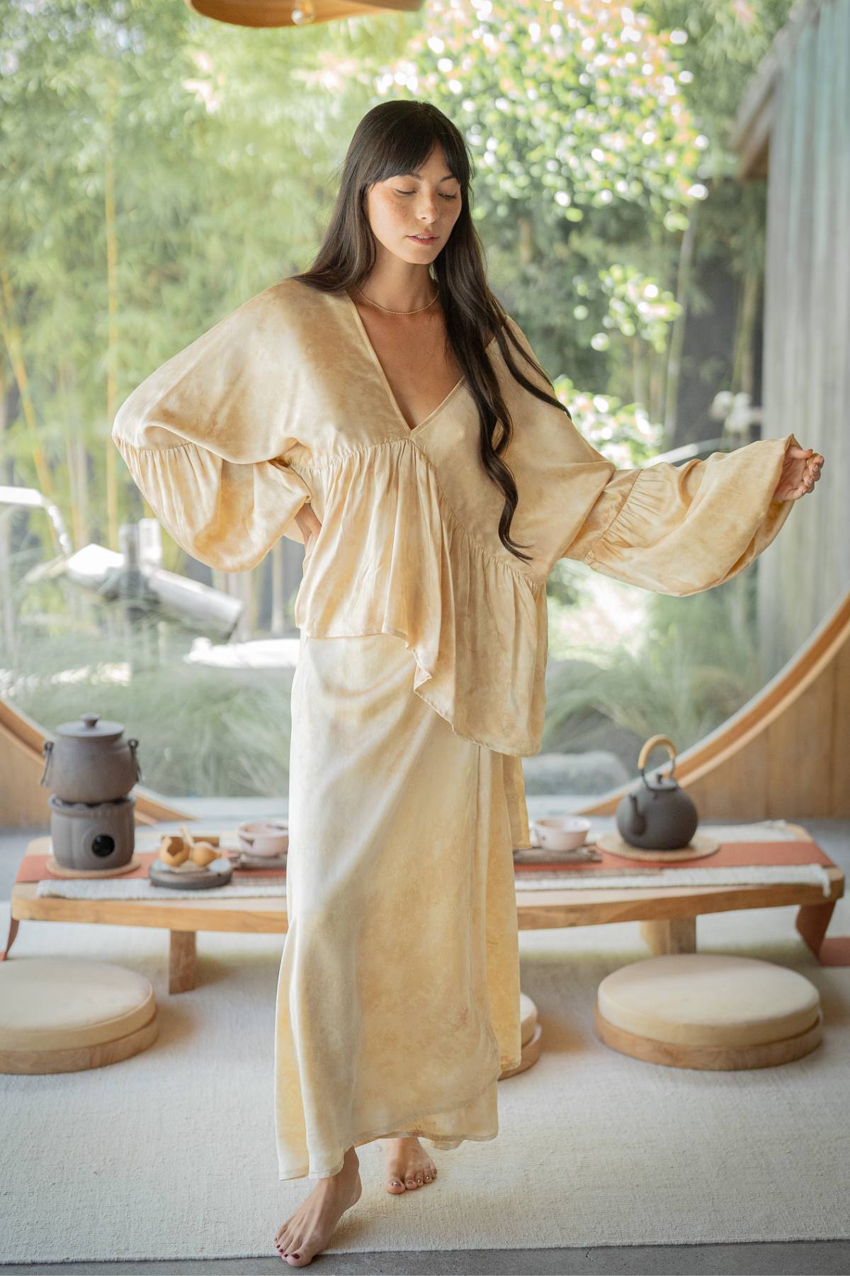In a tranquil indoor setting, a woman with long dark hair stands barefoot, dressed in an elegant Eva Top & Shanti Skirt Set by Myrah Penaloza, crafted from 100% naturally dyed silk and made to order. Behind her is a rustic table adorned with a tea set, while through the large circular window, vibrant greenery is visible.