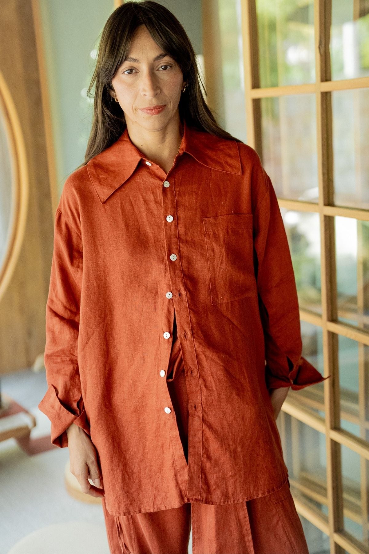 A person with long, dark hair stands indoors wearing the Sutra Linen Set (Made To Order) by Myrah Penaloza—an oversized, burnt orange button-up shirt paired with matching pants. They have a calm expression and stand near a wooden partition with glass panes, allowing soft natural light to filter in.