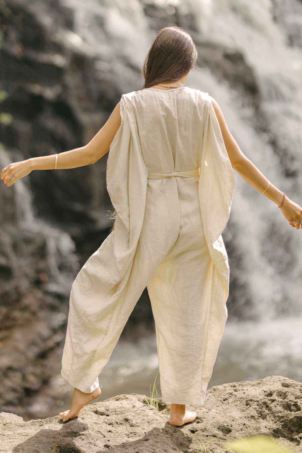 A person with long hair, wearing the beige French Linen Kuan Yin Playsuit by Myrah Penaloza, which features an adjustable waist tie, stands on a rock facing a waterfall. Their arms are slightly outstretched, and they appear to be barefoot, enjoying the natural surroundings.