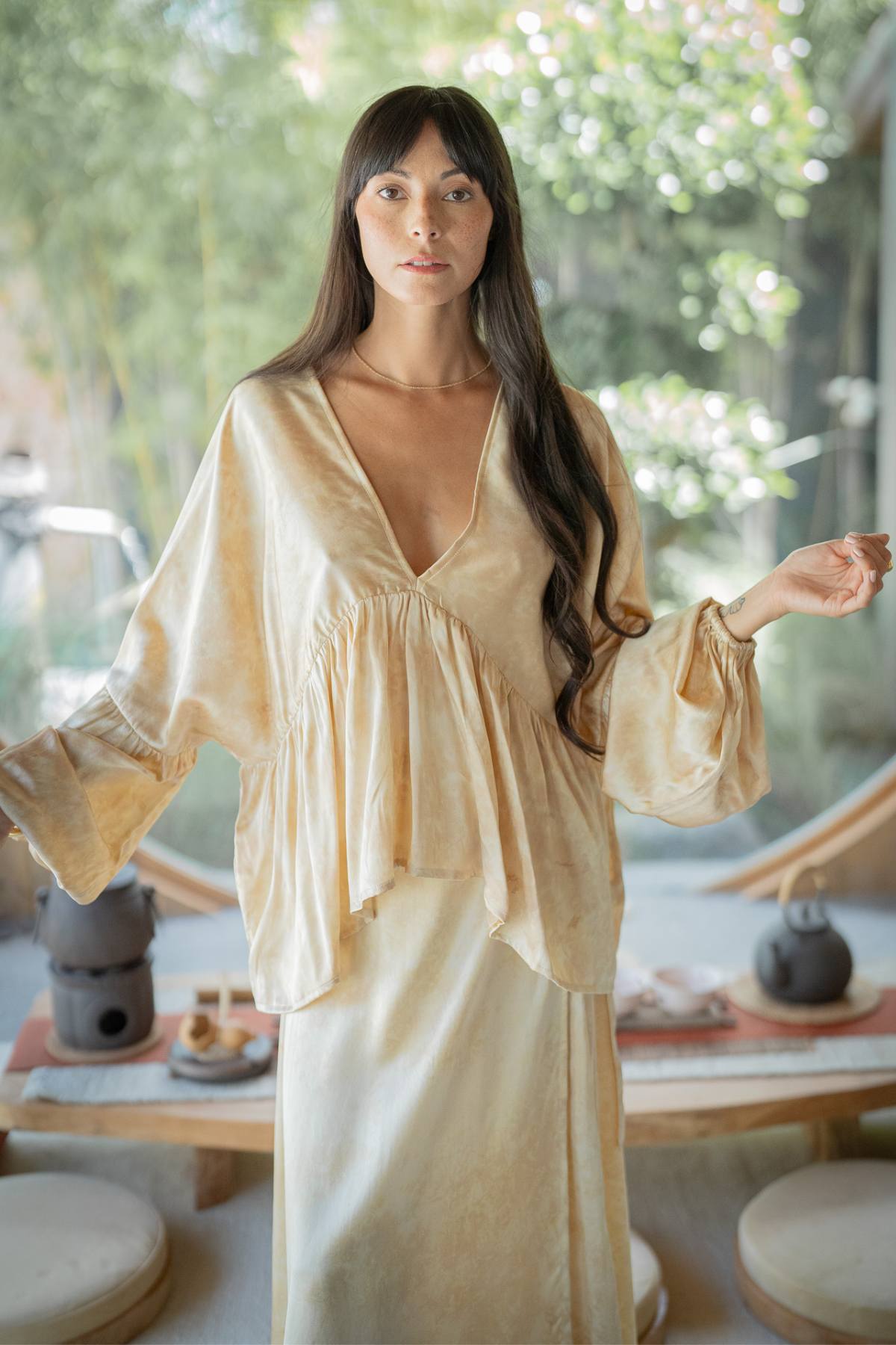 A woman models the Eva Top & Shanti Skirt Set from Myrah Penaloza, crafted from 100% naturally dyed silk. She stands indoors with her long dark hair cascading down as she holds her arms slightly out to the sides. In the background, a teapot rests on a low table, while the blurred greenery beyond adds a serene touch.