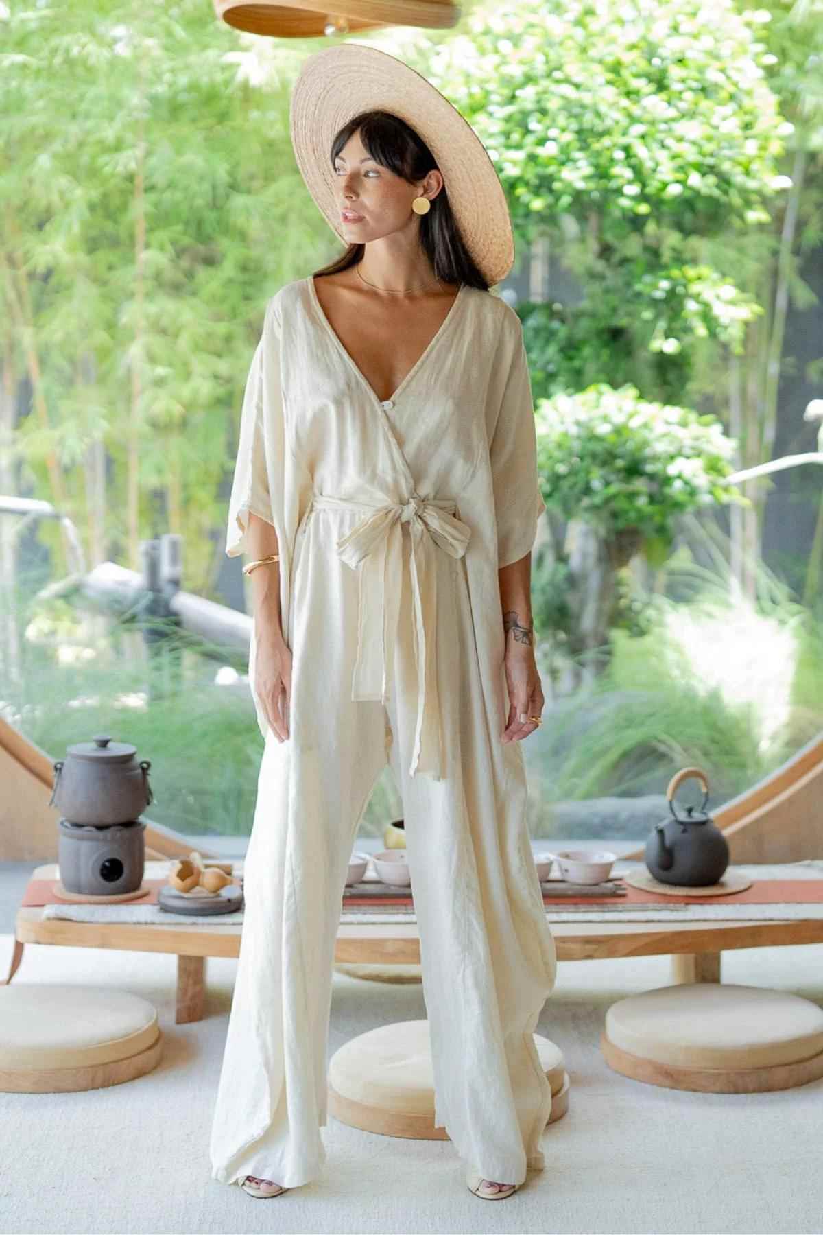 A woman stands indoors wearing a light, flowing, cream-colored stone-washed linen River Playsuit from Myrah Penaloza with a deep neckline and a wide-brimmed straw hat. She is in a tranquil setting with lush green plants visible in the background and a wooden tea setup in the foreground.