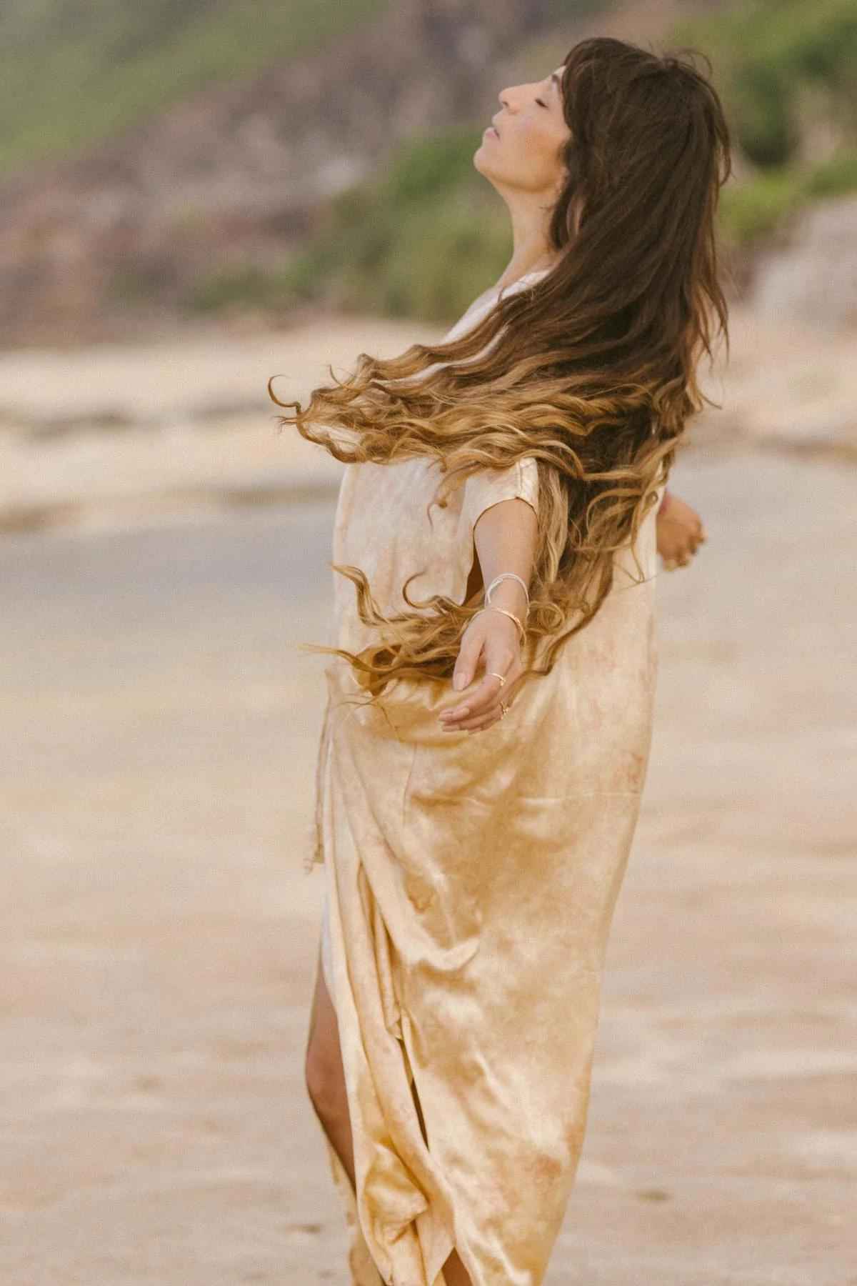A person with long, flowing brown hair wearing a loose, Silk Virgo Kaftan Botanically Dyed by Myrah Penaloza stands on a beach with arms outstretched, eyes closed, seemingly enjoying the breeze. The sandy shore and blurred greenery in the background suggest a serene, lifestyle setting.