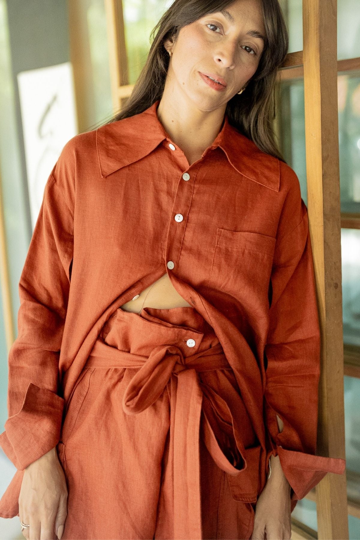 A woman with long dark hair is wearing a loose-fitting, rust-colored Sutra Linen Set by Myrah Penaloza, consisting of a button-up shirt and matching high-waisted pants with a tied belt. She stands against a wooden and glass door with a relaxed expression, showcasing her sustainable look.