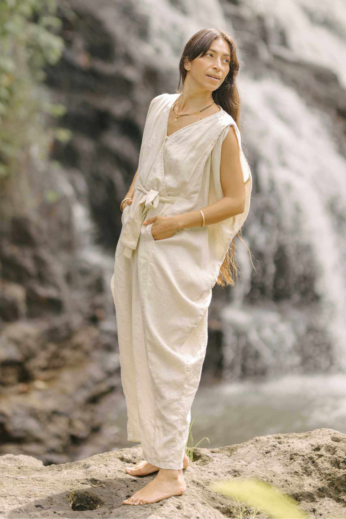 A person with long hair stands barefoot on a rock near a waterfall, dressed in the Myrah Penaloza French Linen Kuan Yin Playsuit. The light-colored, sleeveless playsuit features an adjustable waist tie. They gaze to the side with a calm expression, surrounded by cascading water and lush greenery.