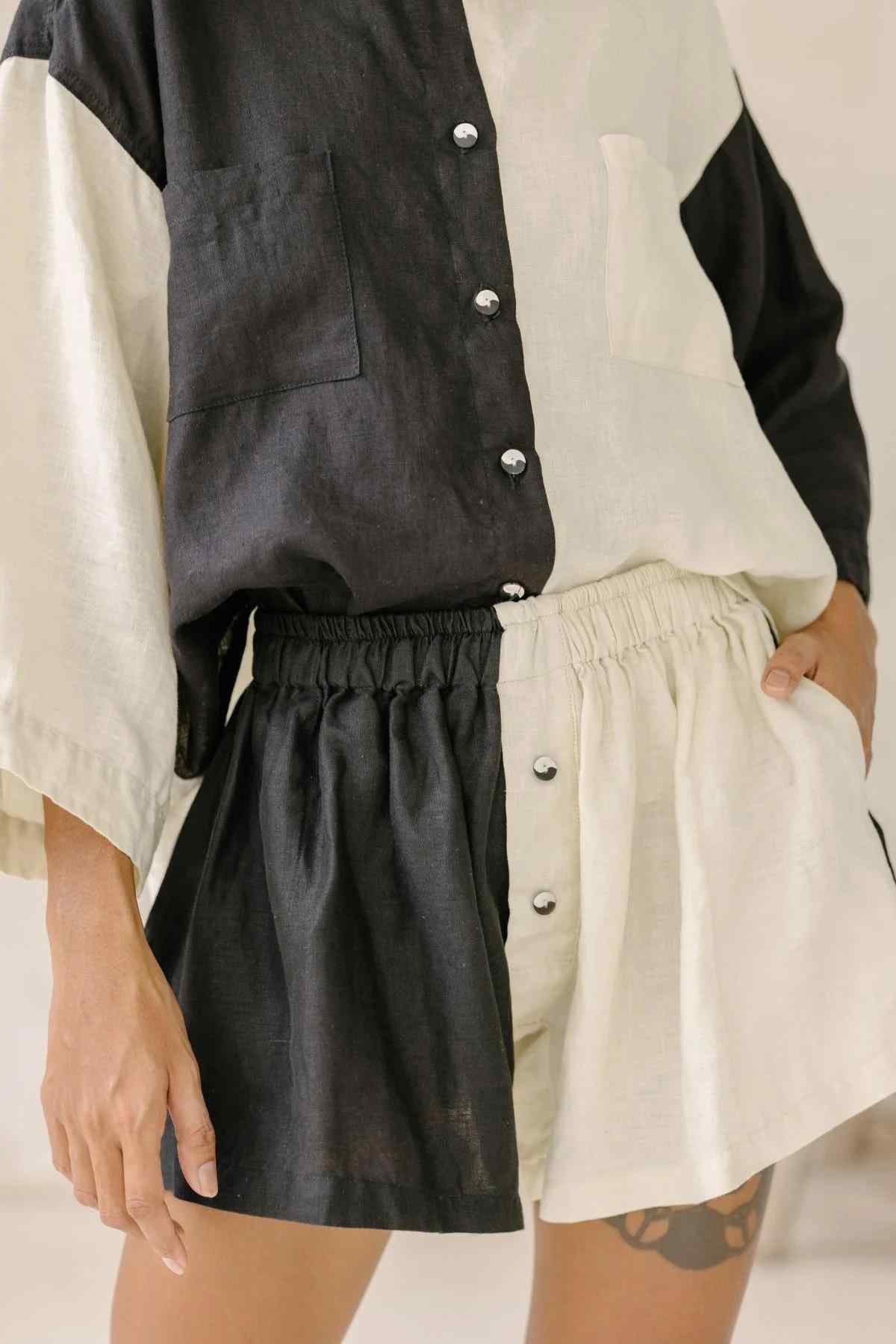 A person models the Yin Yang Nidra Button Down Set (Made To Order) by Myrah Penaloza, featuring a relaxed silhouette with a half-black, half-cream-colored linen button-down top and matching shorts with an elastic waistband. The left side of the outfit is black, while the right side is cream-colored. One hand rests on their hip, revealing a tattoo on their thigh.