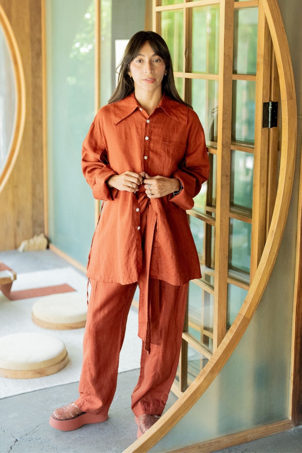 A person with long brown hair stands indoors near a circular doorway, wearing a loose-fitting rust-colored button-up shirt and matching pants from the Sutra Linen Set by Myrah Penaloza. With their hands loosely clasped in front, they appear calm and relaxed. The background features soft lighting and wooden decor, which highlights their sustainable fashion choice with the Made To Order Sutra Linen Set.