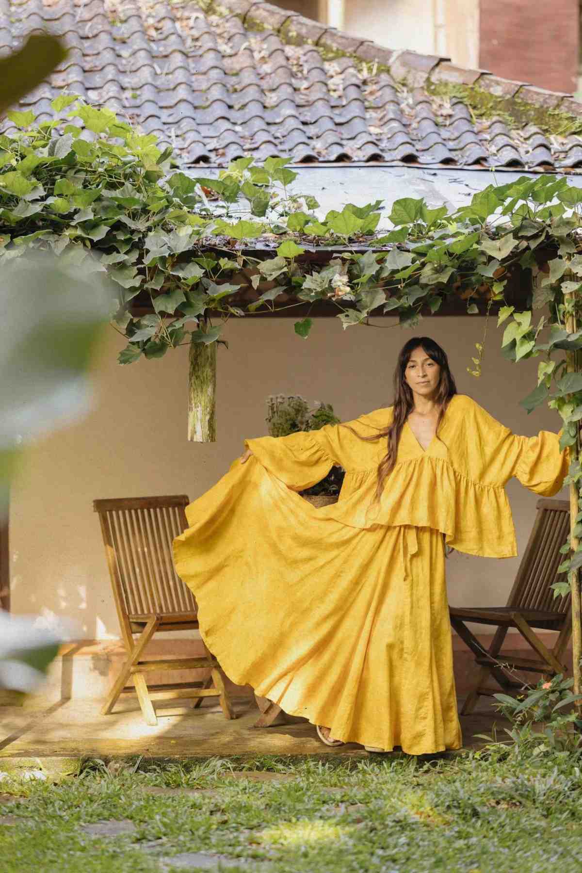 A woman stands outdoors in a garden area near a small, rustic building with a tiled roof. She is wearing the Linen Eva Top & Orisa Dance Skirt Set by Myrah Penaloza, featuring a flowing, bright yellow Orisa Dance Skirt with wide sleeves. Holding the ends of the skirt out, she displays its full length amidst the surrounding greenery.
