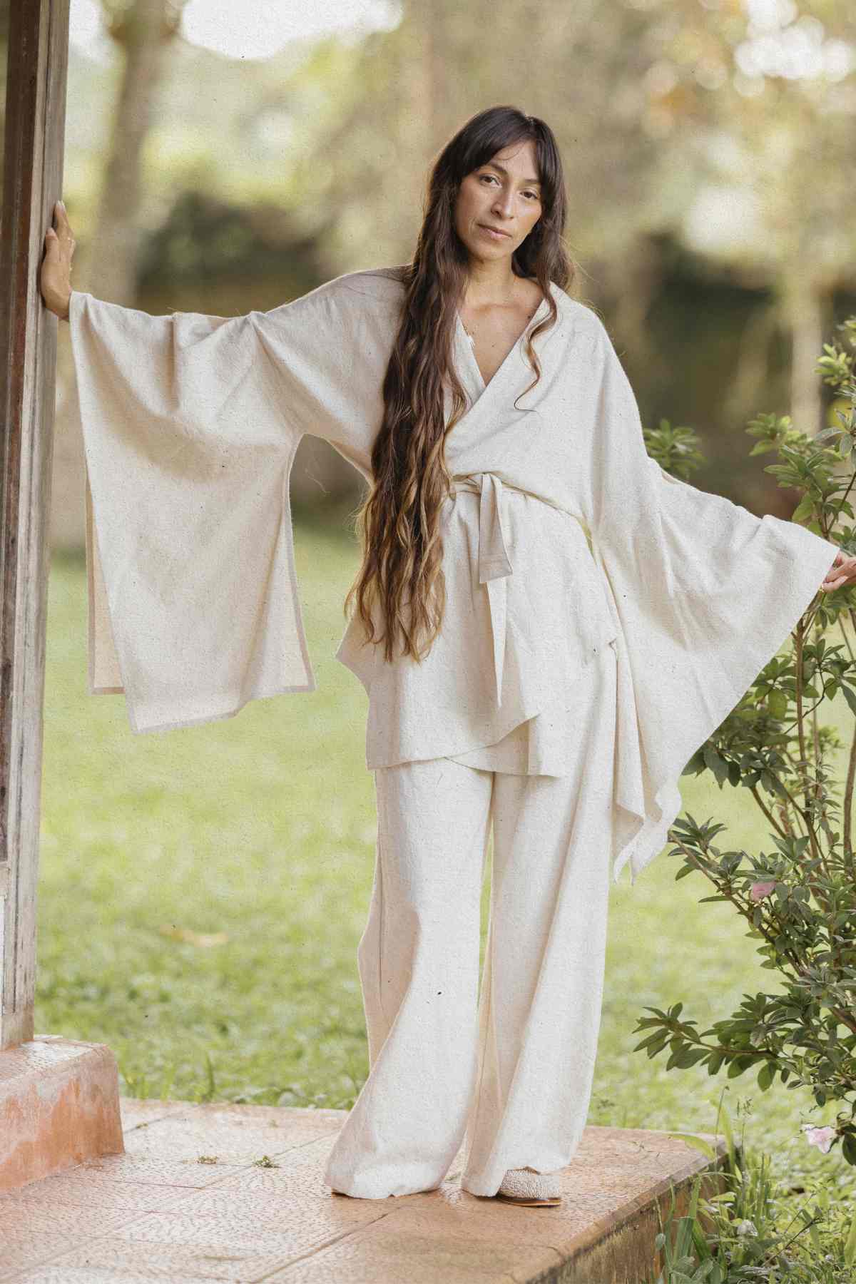 A woman with long hair stands outdoors on a tiled surface, leaning against a column. She is wearing the Dewi Set (3 of a kind) - Pre-order by Myrah Penaloza, consisting of a loose, light-colored outfit made of hypoallergenic linen cotton crepe with wide sleeves and pants. The background is blurred but appears to be a garden with greenery.