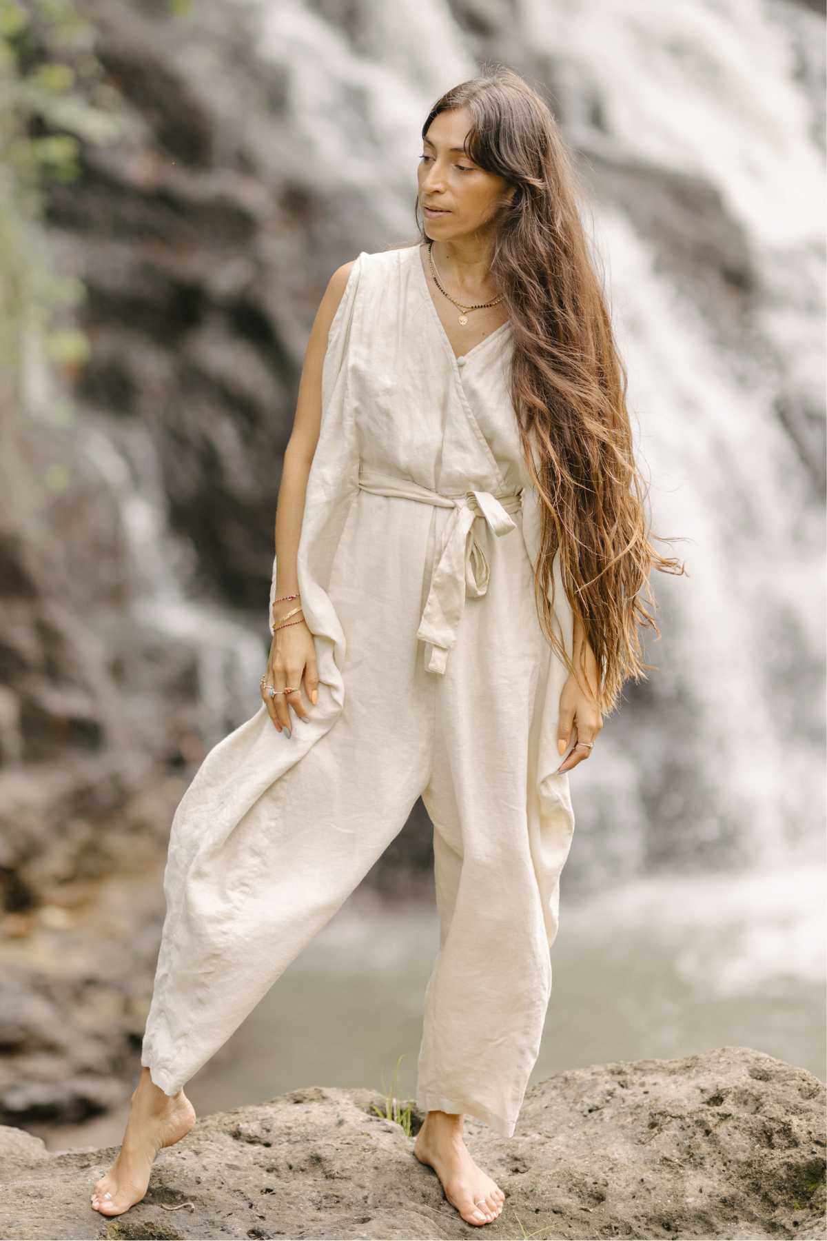A woman with long, flowing hair stands on a rock in front of a waterfall. She is wearing the loose, stonewashed French Linen Kuan Yin Playsuit by Myrah Penaloza, accentuated with an adjustable waist tie. She looks thoughtfully into the distance with one hand resting on her hip and the other hanging by her side.